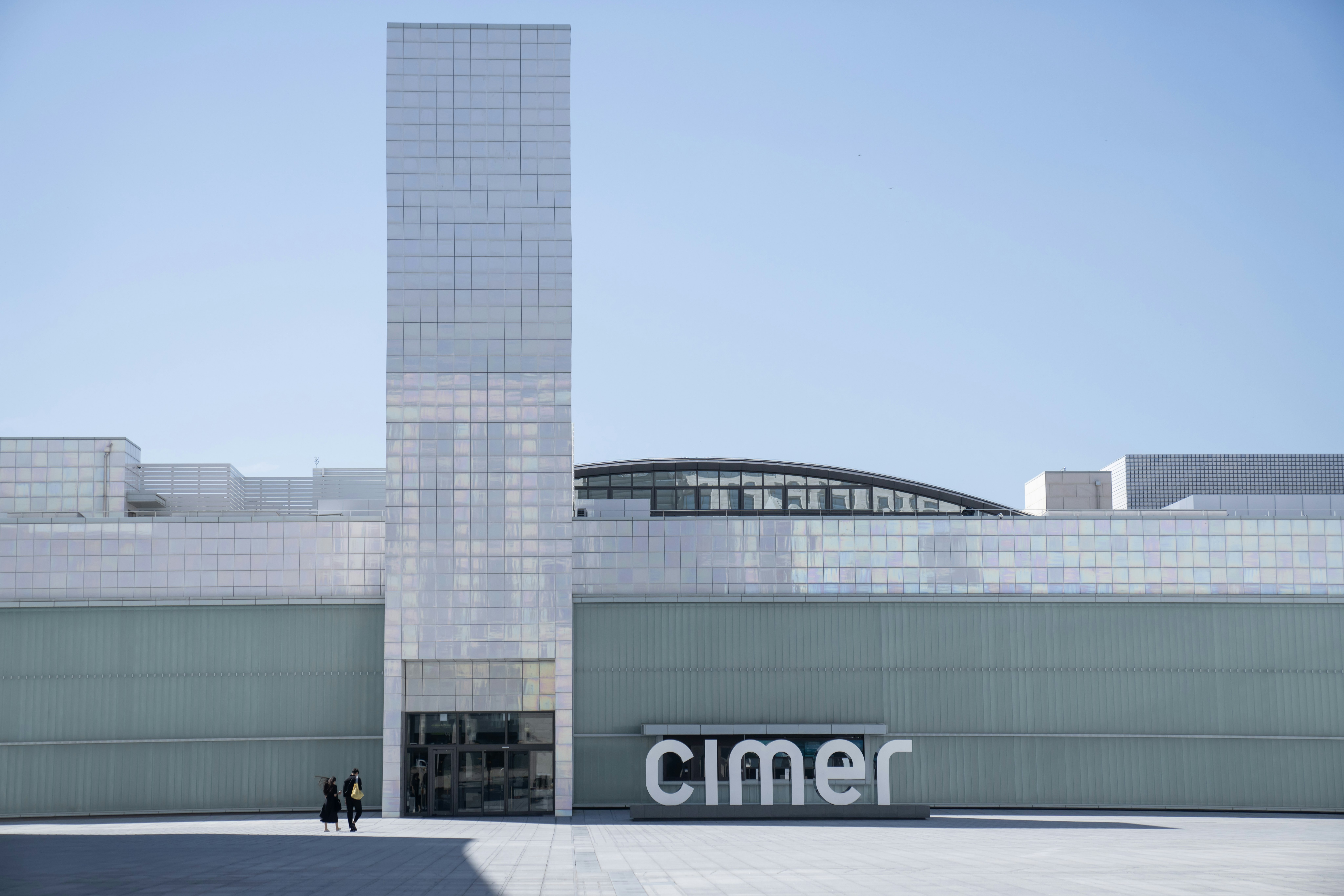 Two people walk towards a vast tiled building with a huge central rectangular tower and a sign that reads "Cimer".