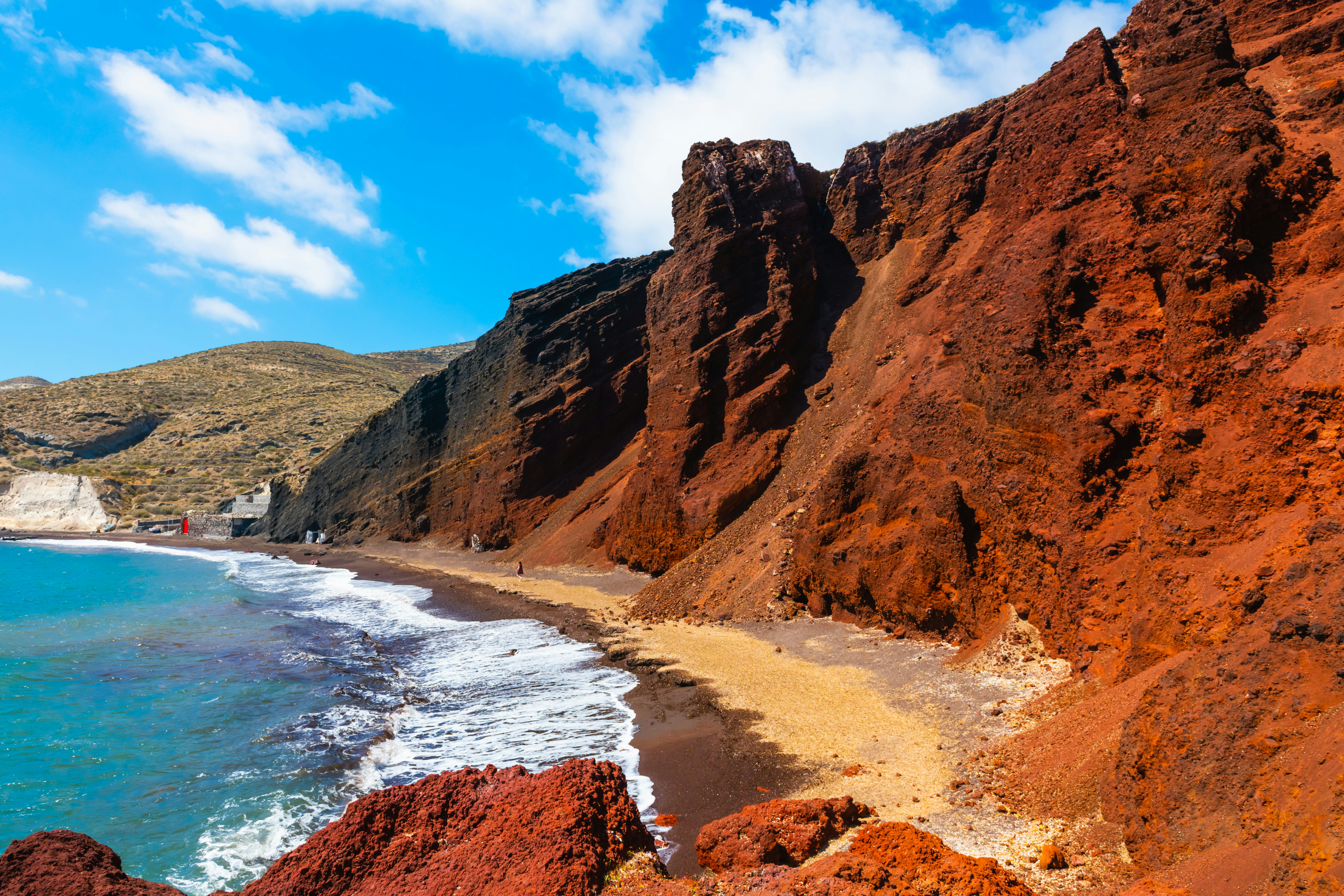 Red volcanic cliffs along a blue sea under light blue skies with thin clouds