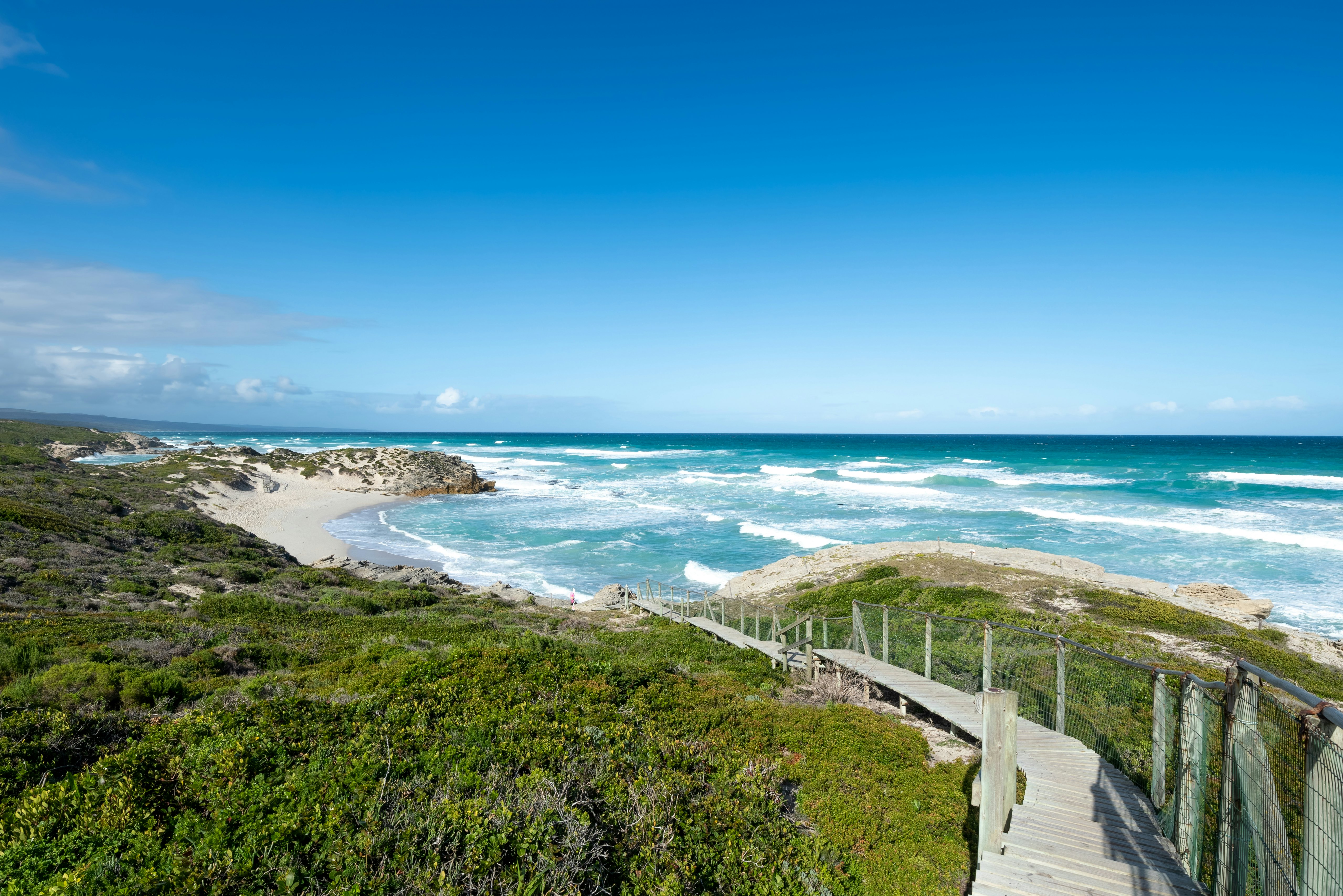 A boardwalk over shrubs and grassland leads down to a sandy beach being pounded by the surf.