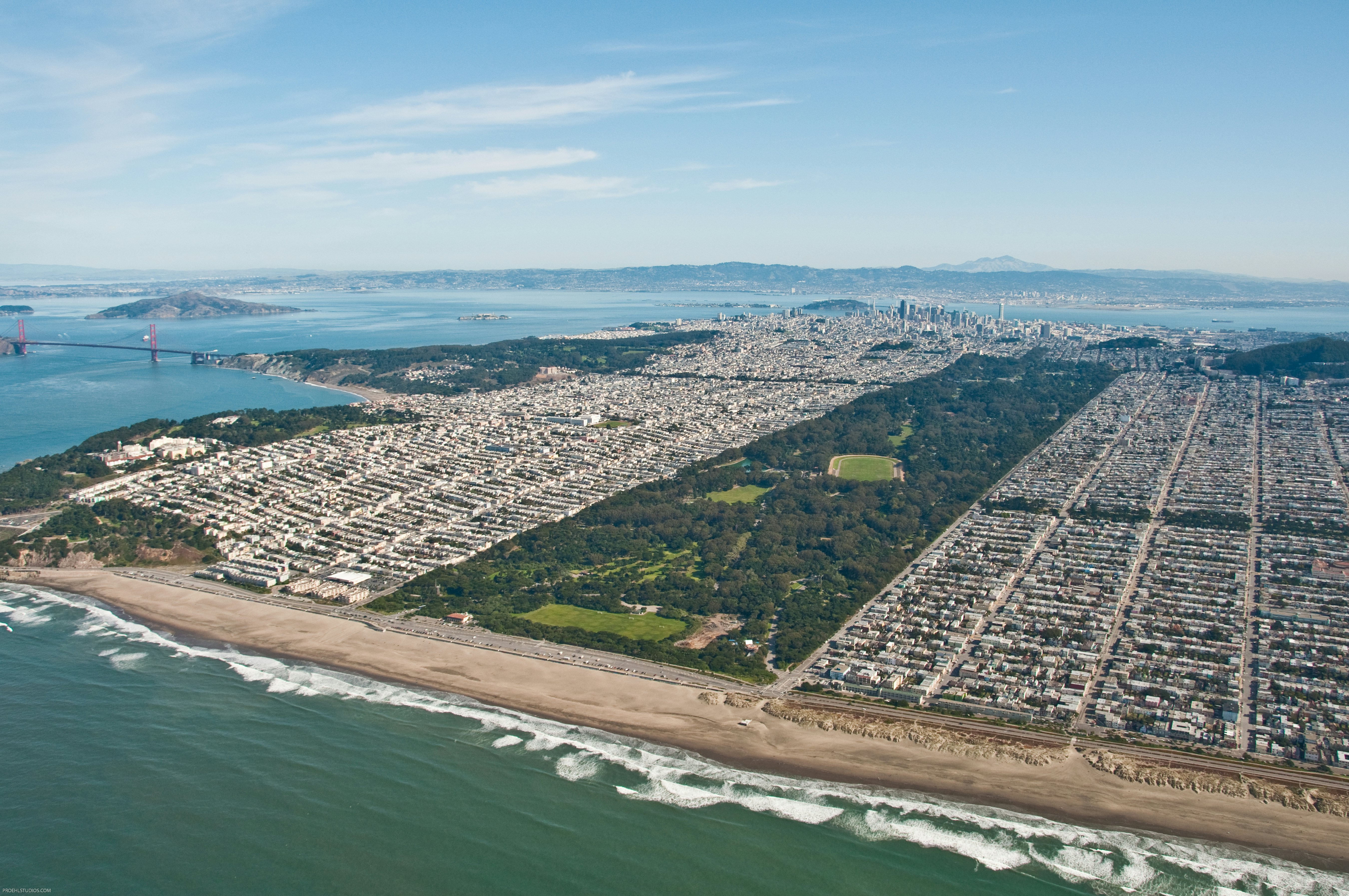 An aerial view of Golden Gate Park bordered by city neighborhoods and surrounded by the ocean with the Golden Gate Bridge and San Francisco Bay in the distance to the left on a party sunny day.