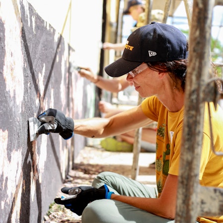 A woman in a yellow T-shirt and black baseball cap helps paint an exterior wall on a sunny day.