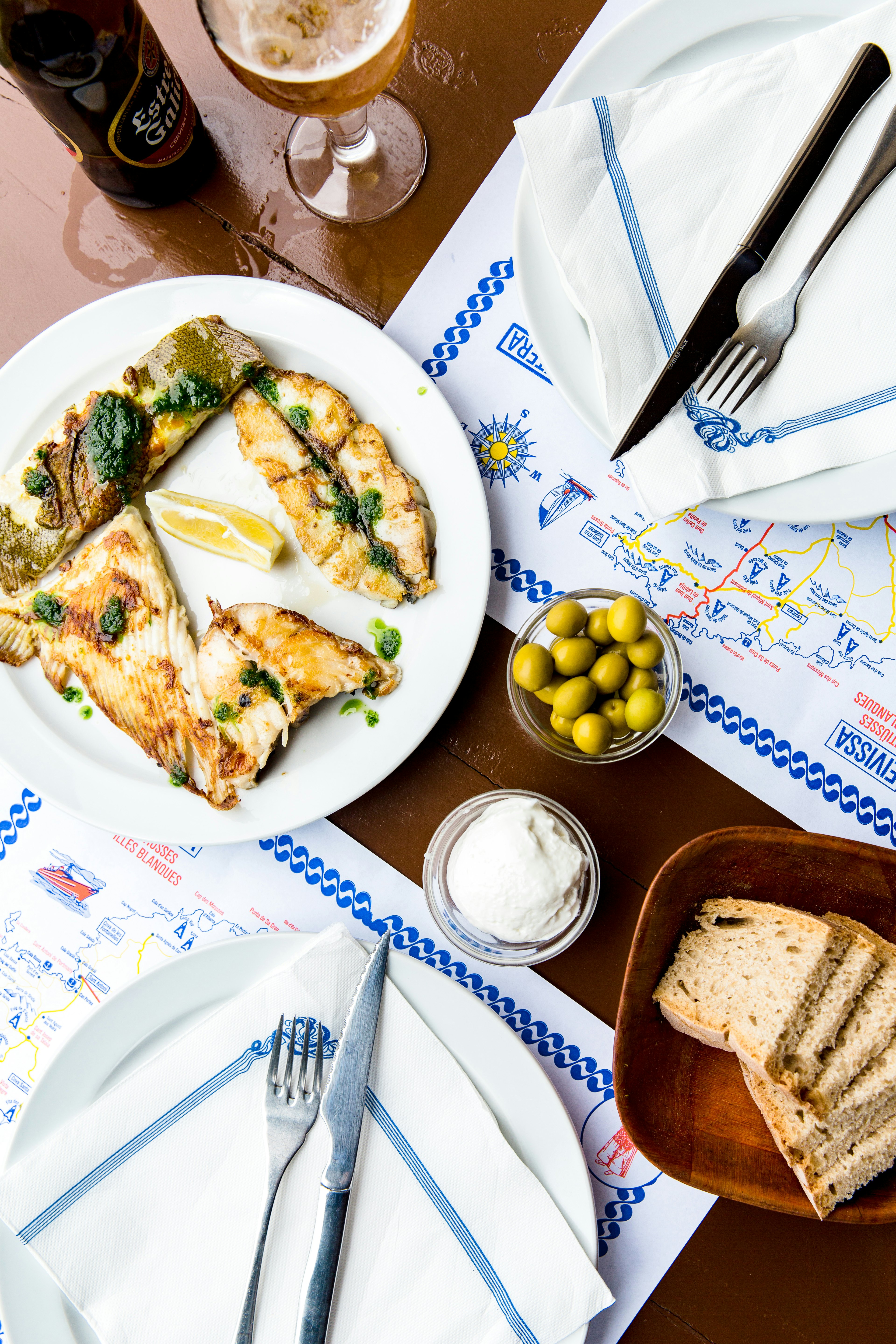 A scene in a restaurant featuring a table with blue and white napkins, a bowl of olives and bread, alongside a plate of grilled fish.