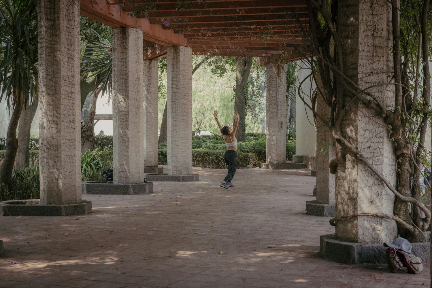 Woman dances with her arms up under a wooden pergola with cement pillars climibing with huge trees and vines. Greenery fills the background