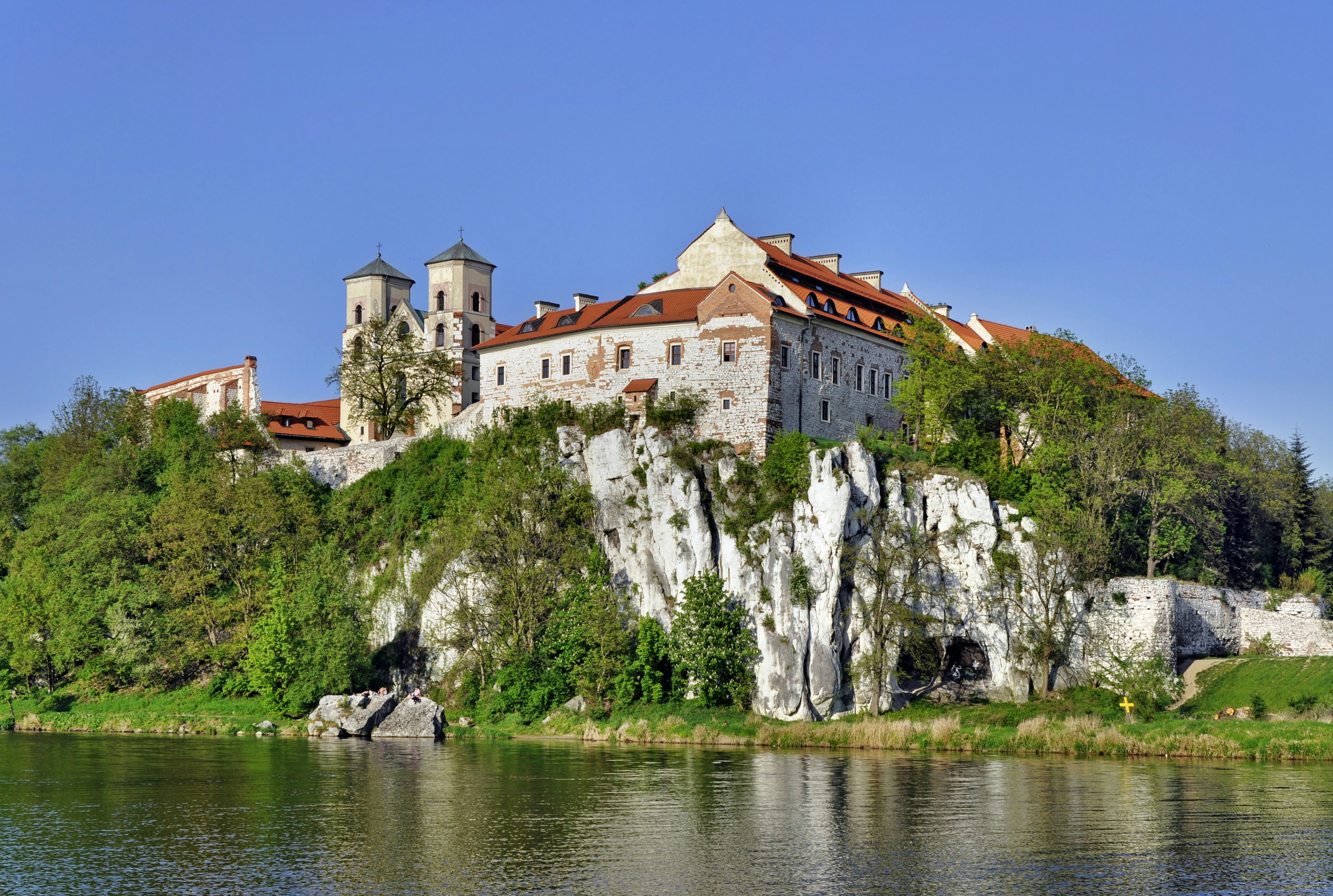 A stone building with a red roof on a rocky hill above a river with grass and trees on the shore.