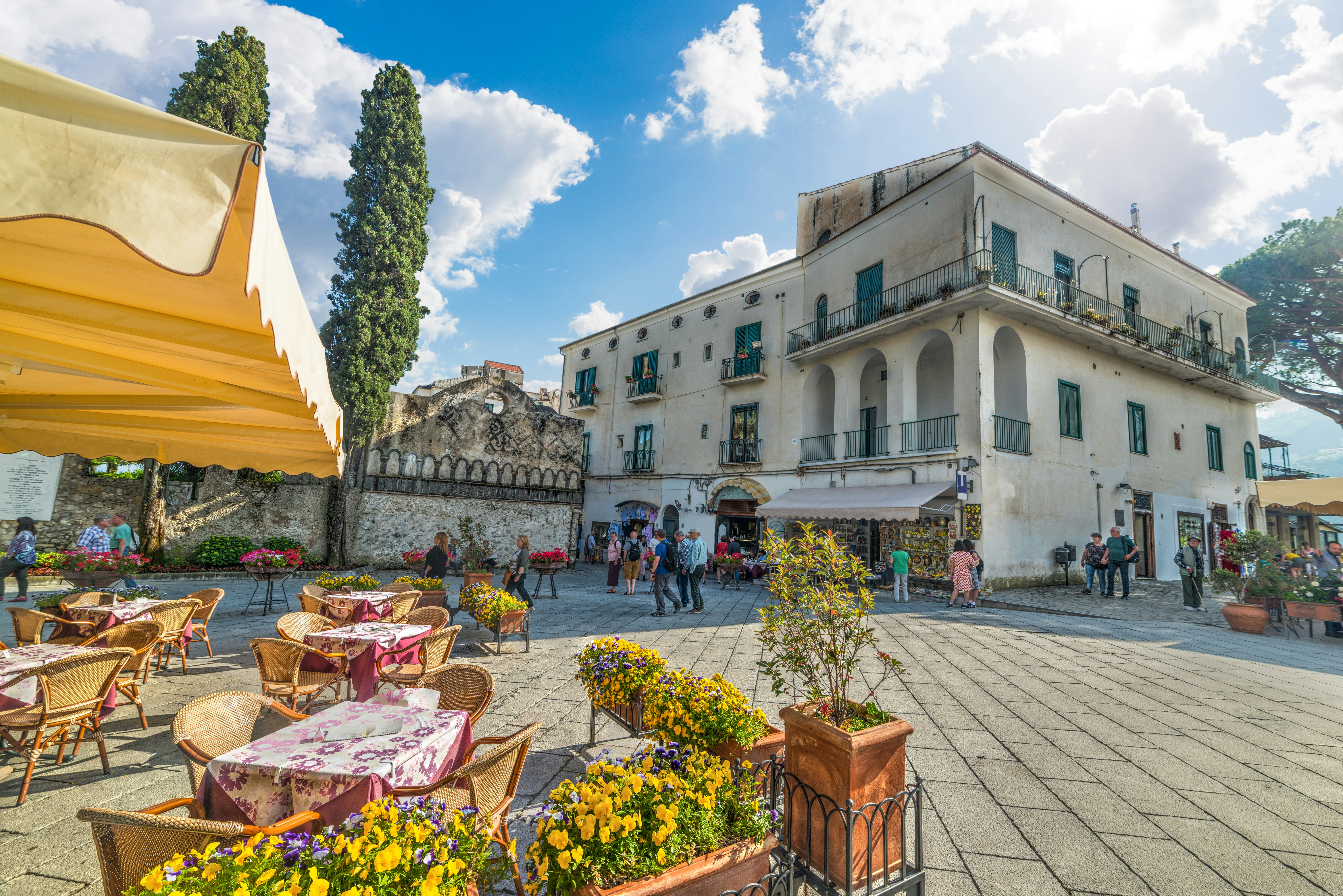 A yellow umbrella provides shade for restaurant tables on the edge of a town square lined with flower boxes.