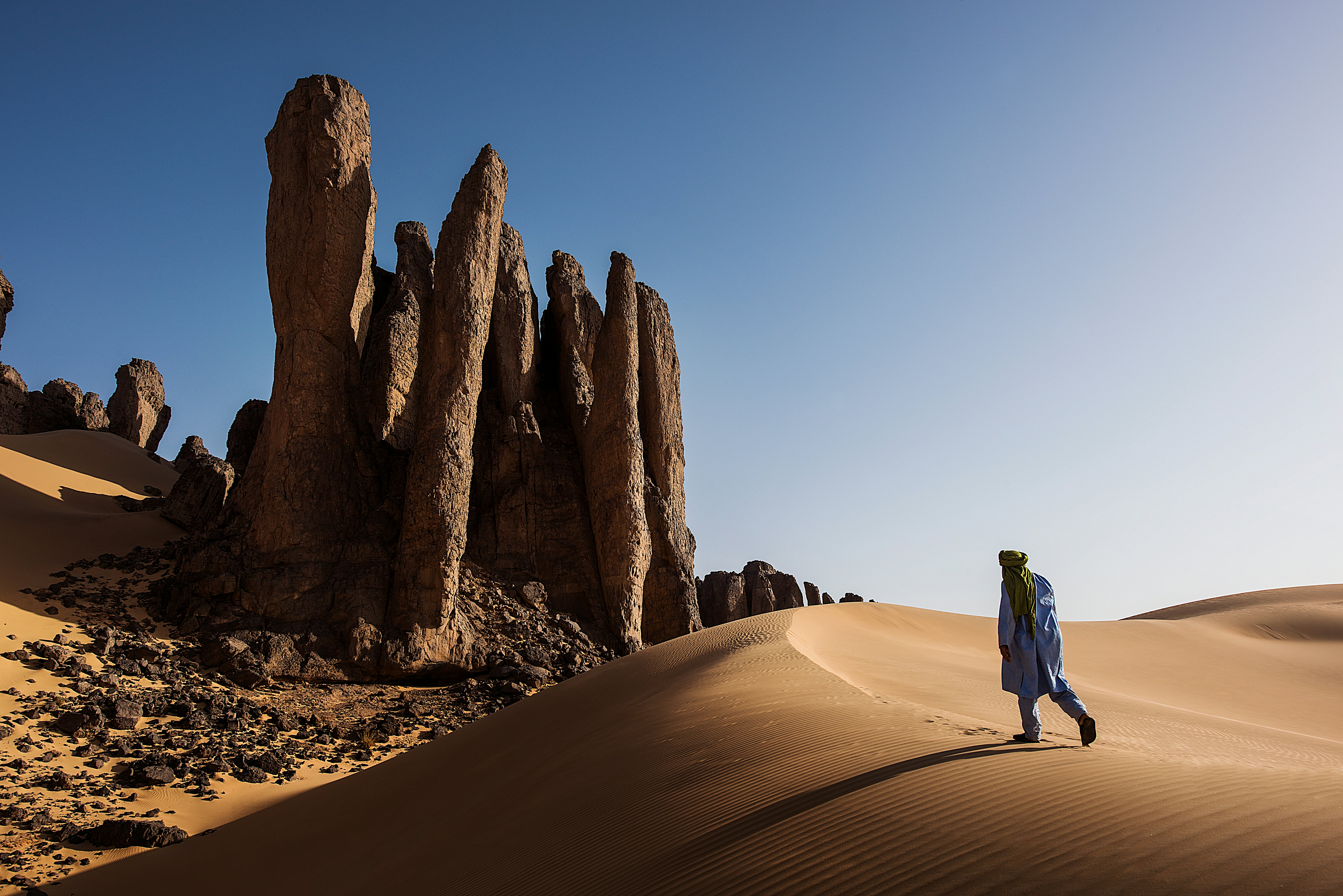 A person walking up a sand dune in Algeria toward rock formations under bright sun and blue skies.