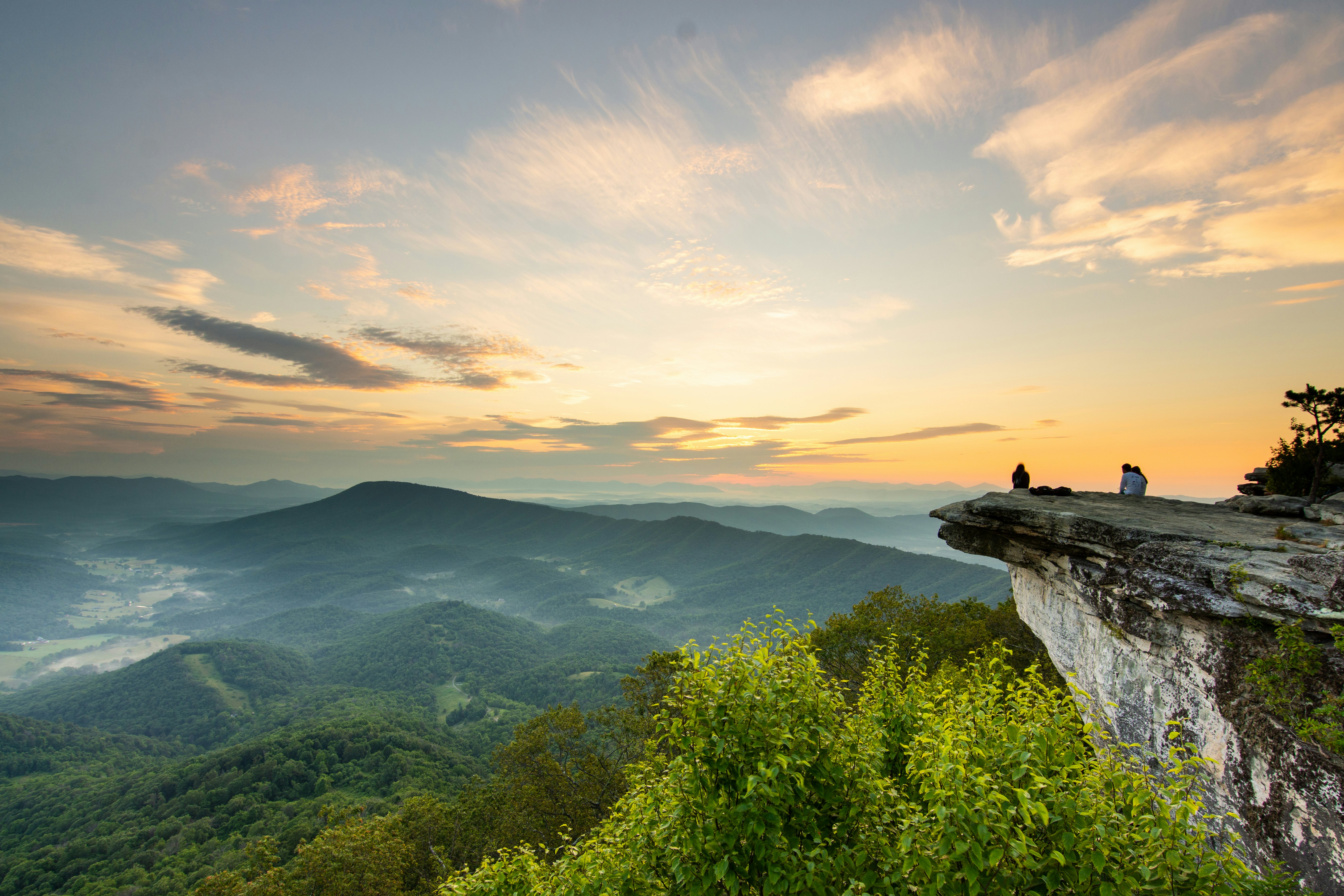 Hikers pause at a viewpoint: a large rocky shelf sticking out over a vast greenery covered valley.