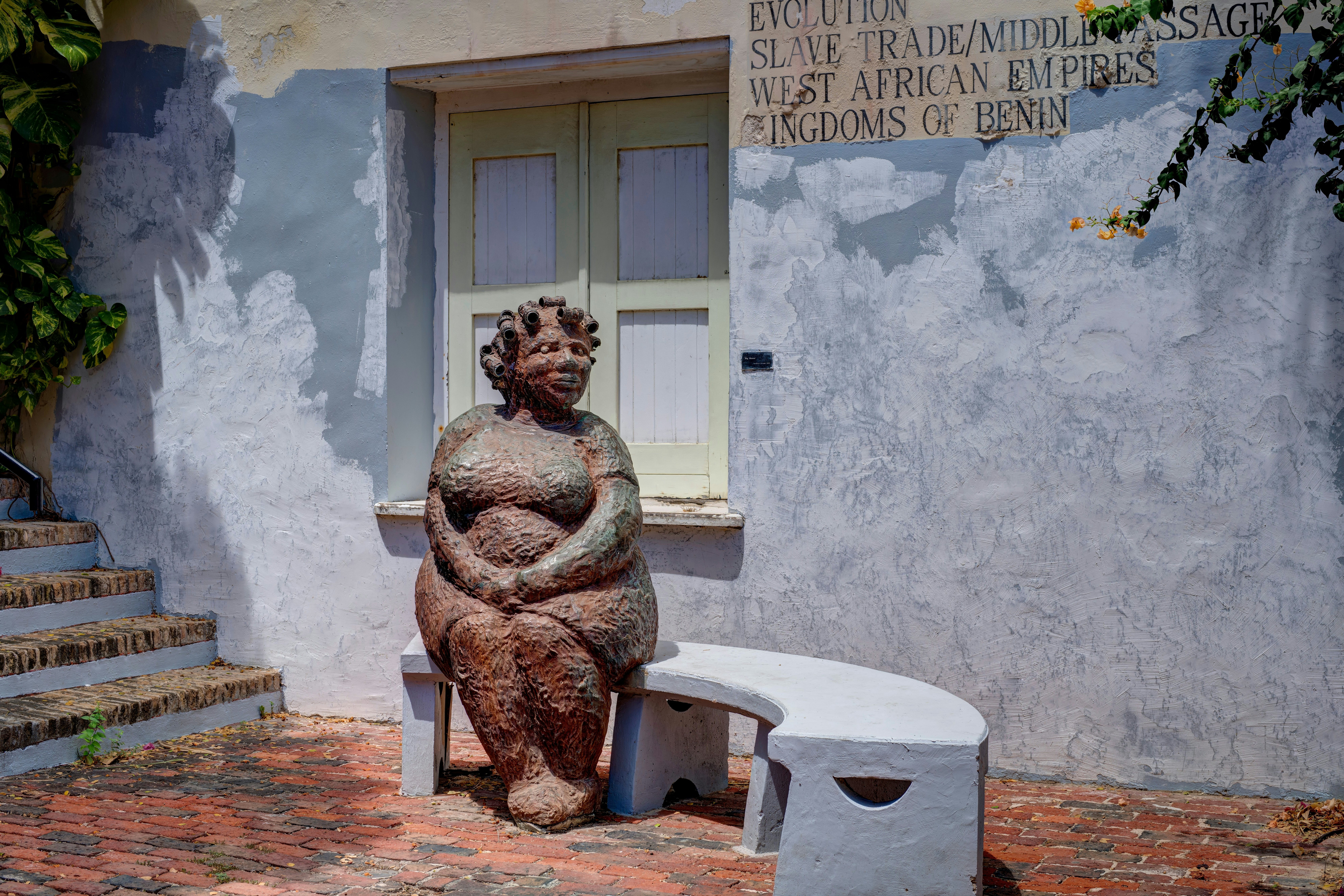 A statue of a woman sat on a bench outside a building with the words "slave trade/middle passage west African Empires Kingdoms of Benin" printed on its pale blue walls.