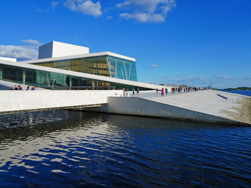 Modern cement and glass-walled building with pier that dips into the surrounding harbor on a sunny day.