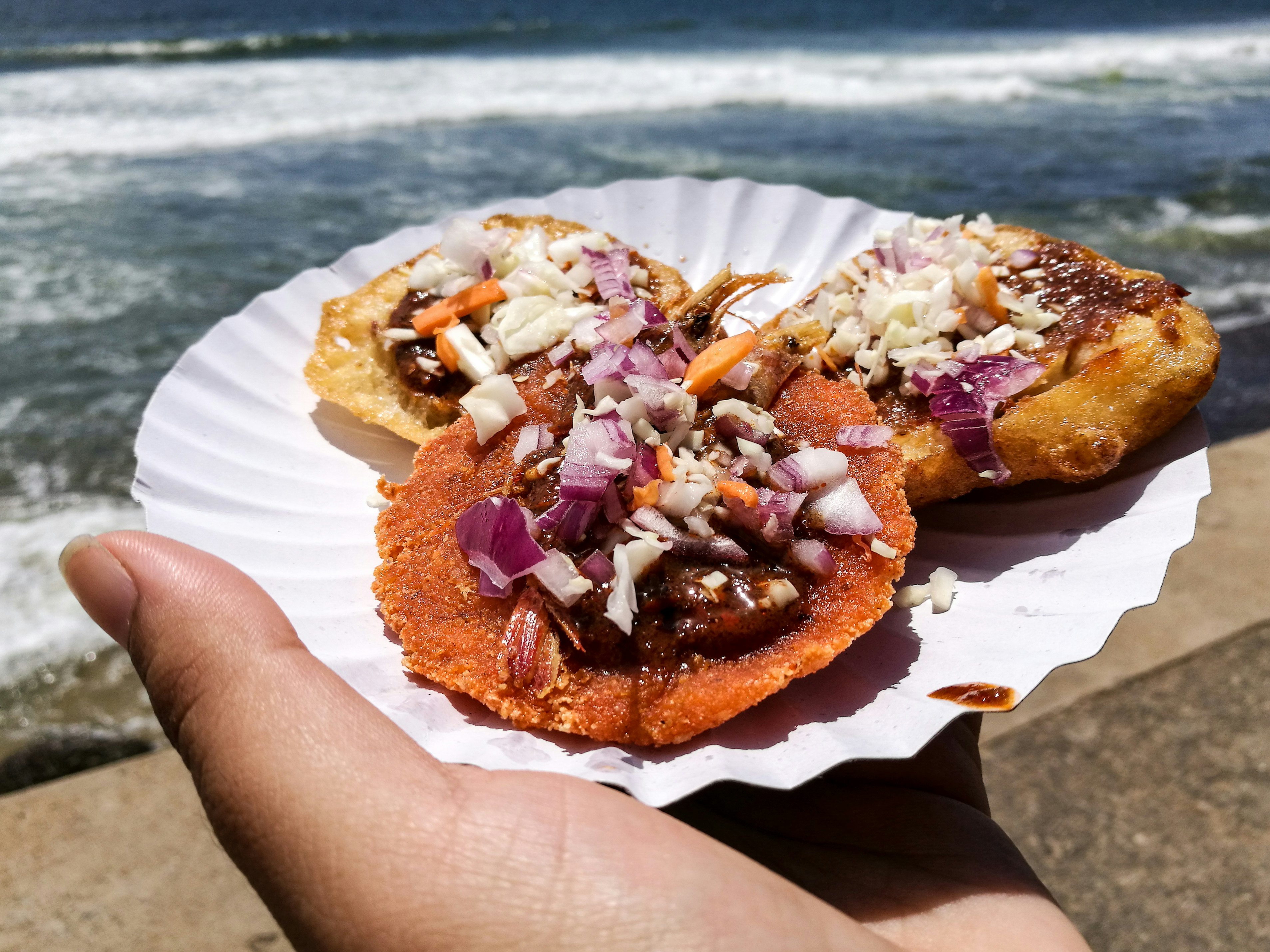 A hand holds out a paper plate loaded with three different pieces of street food on a fried-bread base including onions, vegetables and prawn.