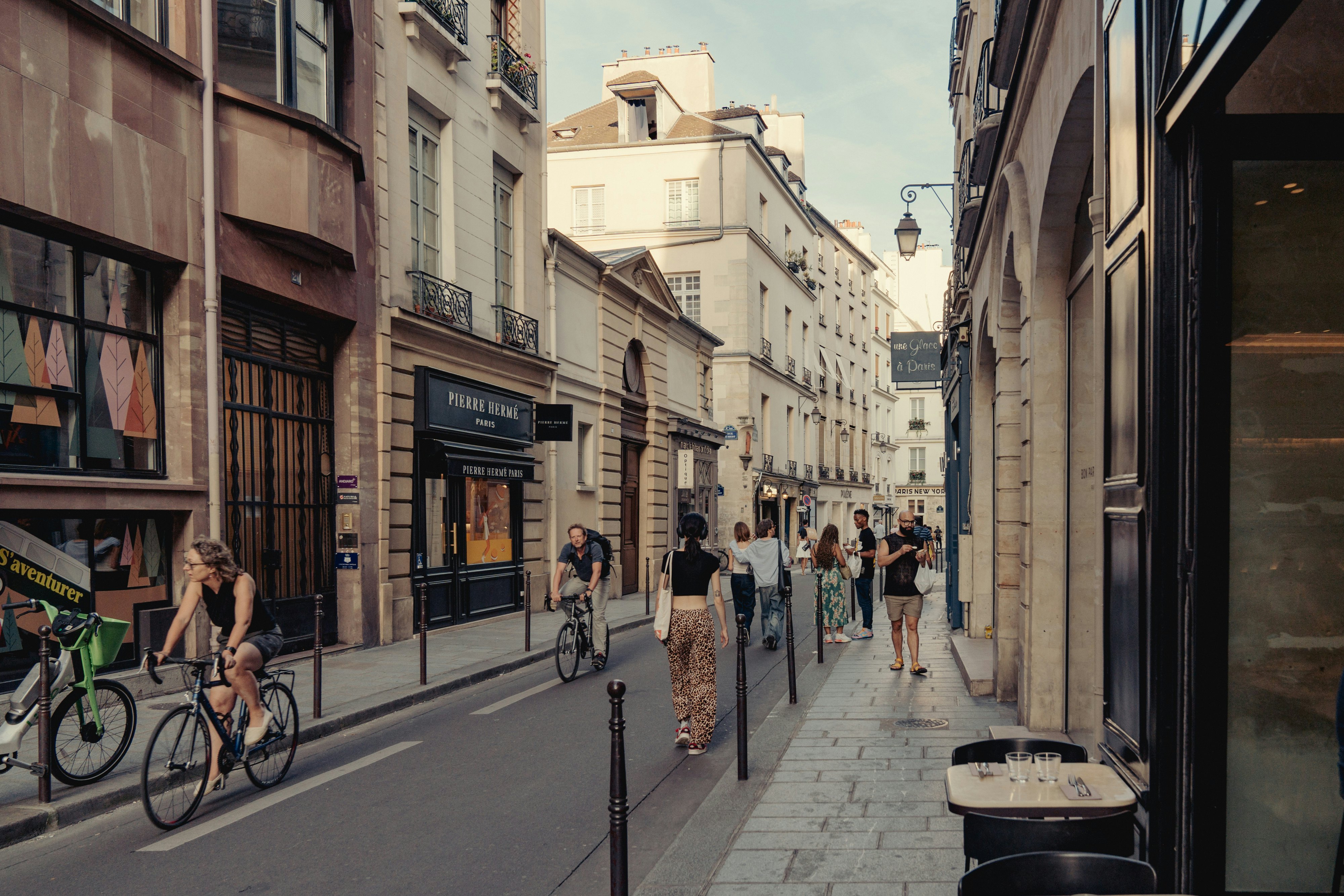A narrow street with cyclists and shoppers passing through. The street is lined with boutiques and cafes.