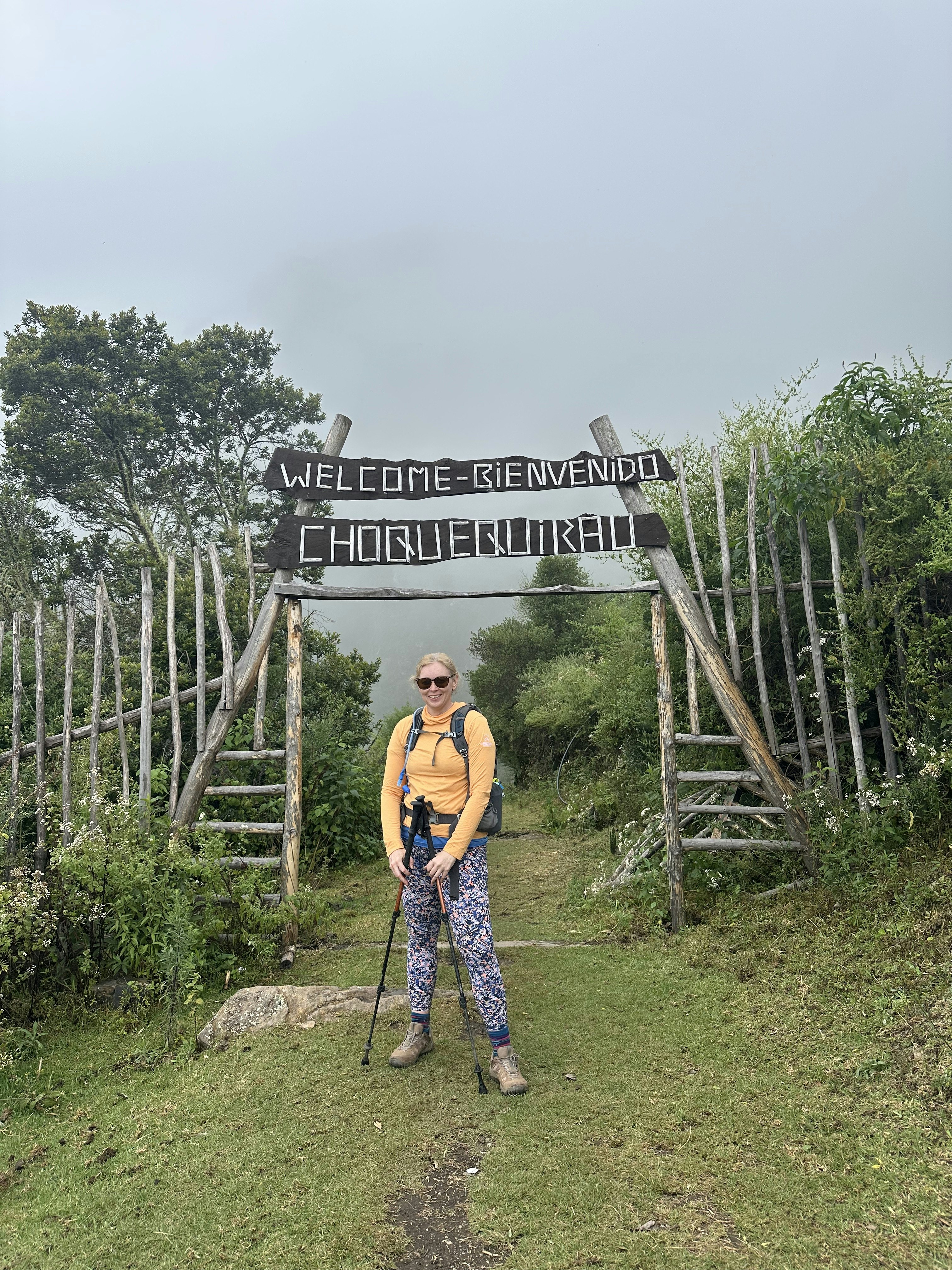 Woman in a yellow top and multicolored pants standing in front of the sign for Choquequirao National Park in Peru. Sign says "Welcome-Bienvenido Choquequiraro."