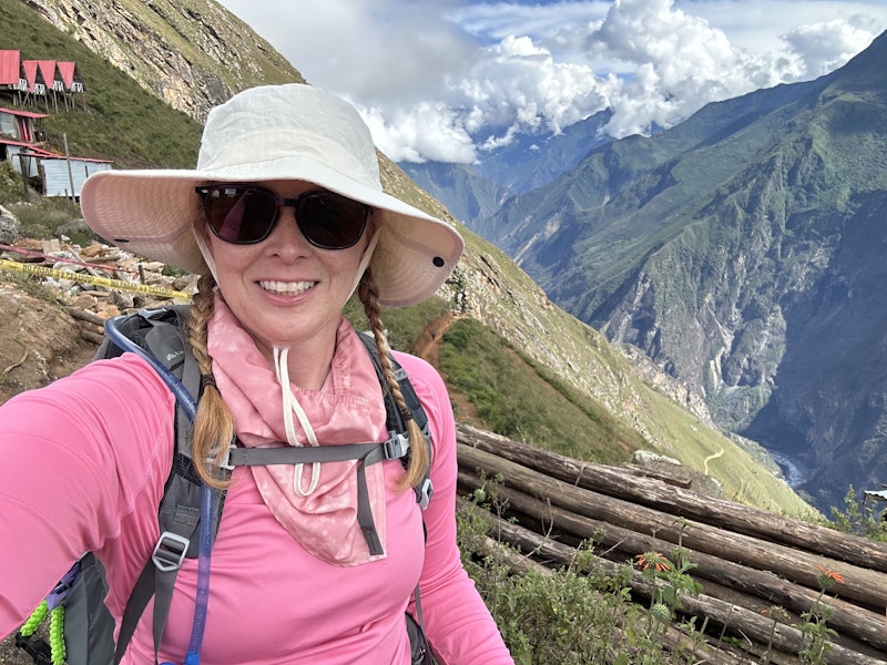 Woman in pink top and white hat and sunglasses smiling with mountainous scenery behind her