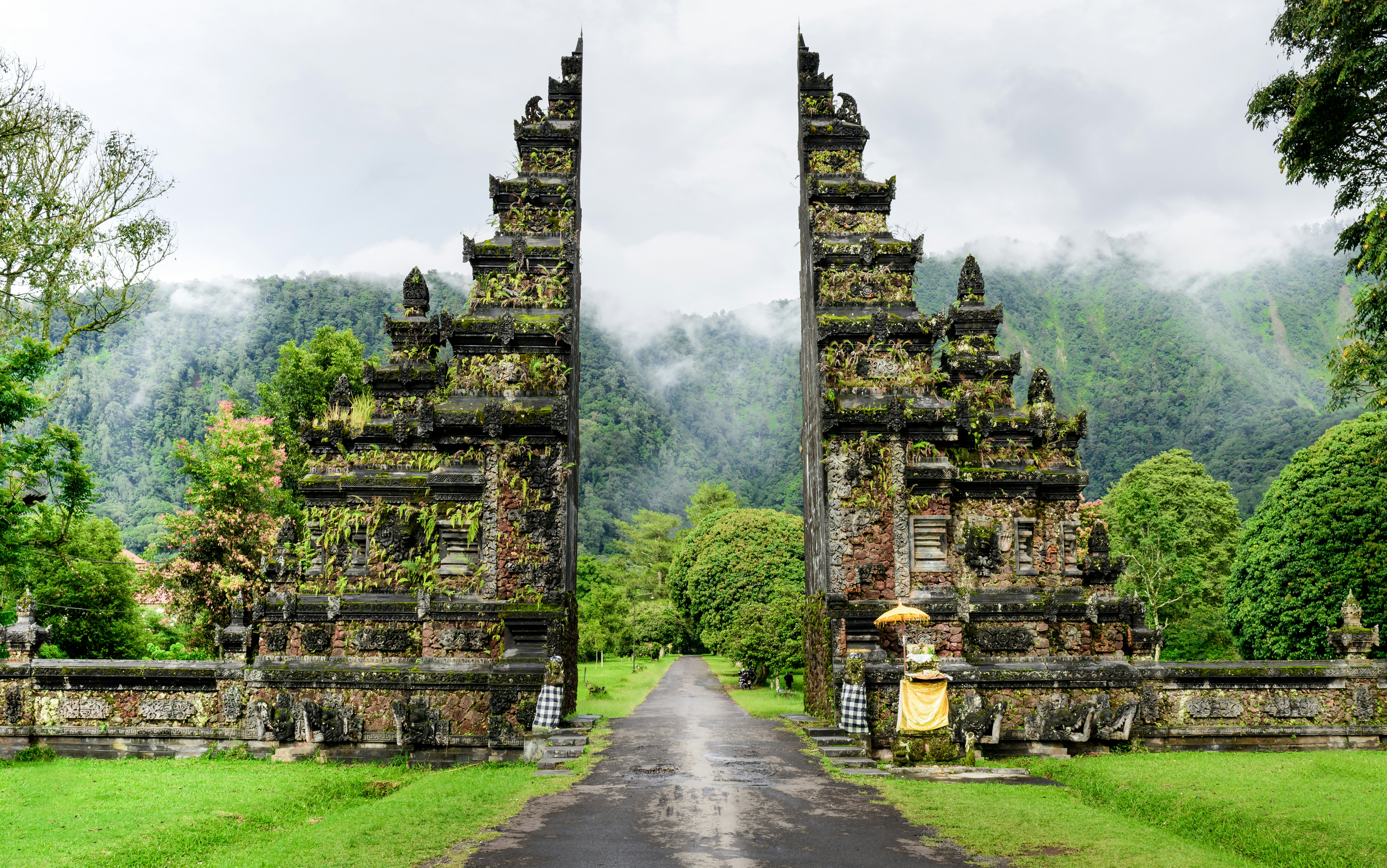 Moss- and fern-covered stone gates beside a concrete path with green grass on either side and misty clouds and a green-treed hill in the distance.