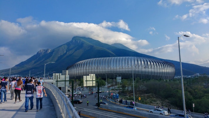 People walk across a bridge toward a round stadium with mountains in the background