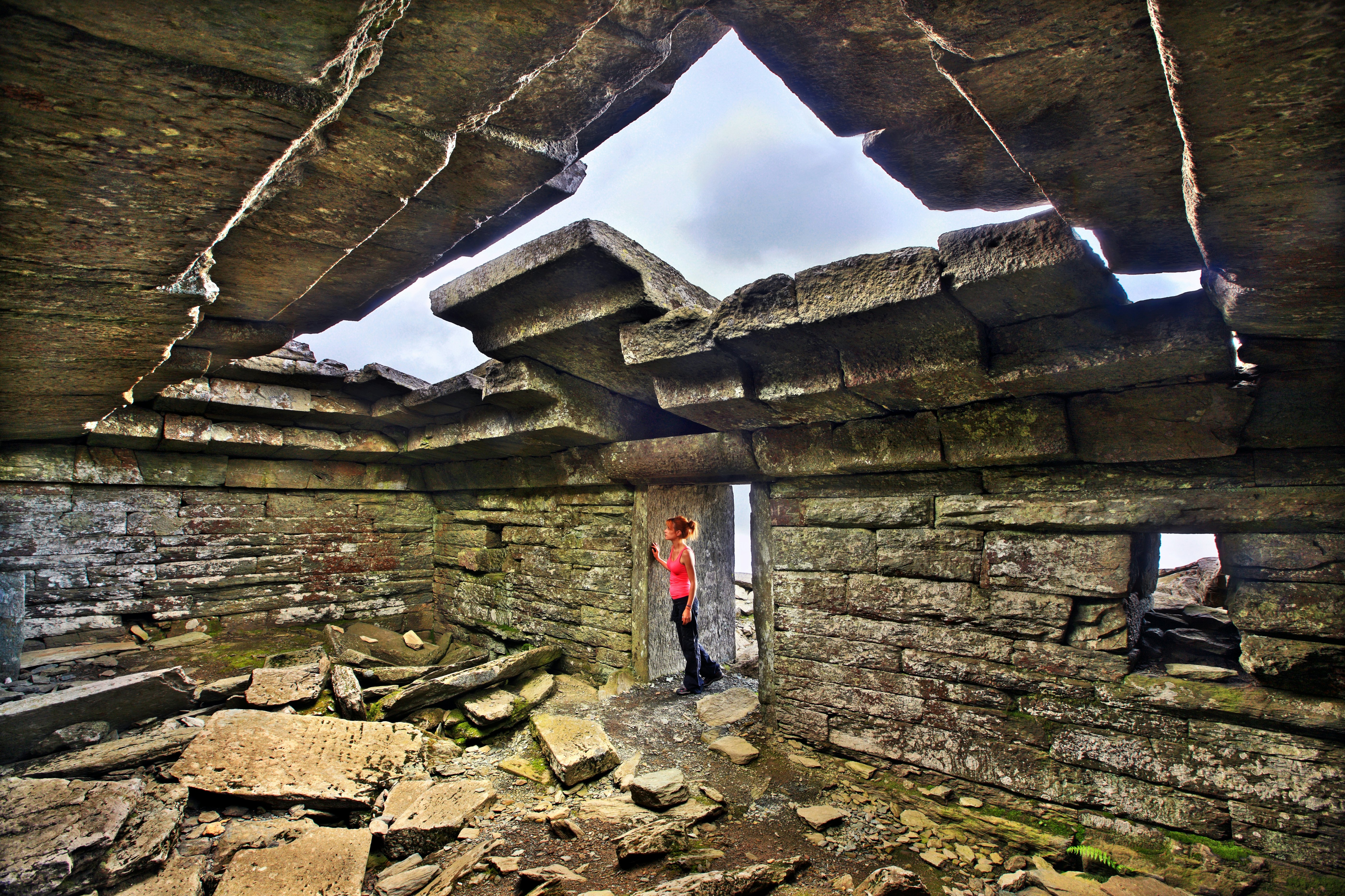 A hiker peers through the doorway of an ancient stone house with crumbling rocks and stones in its center.