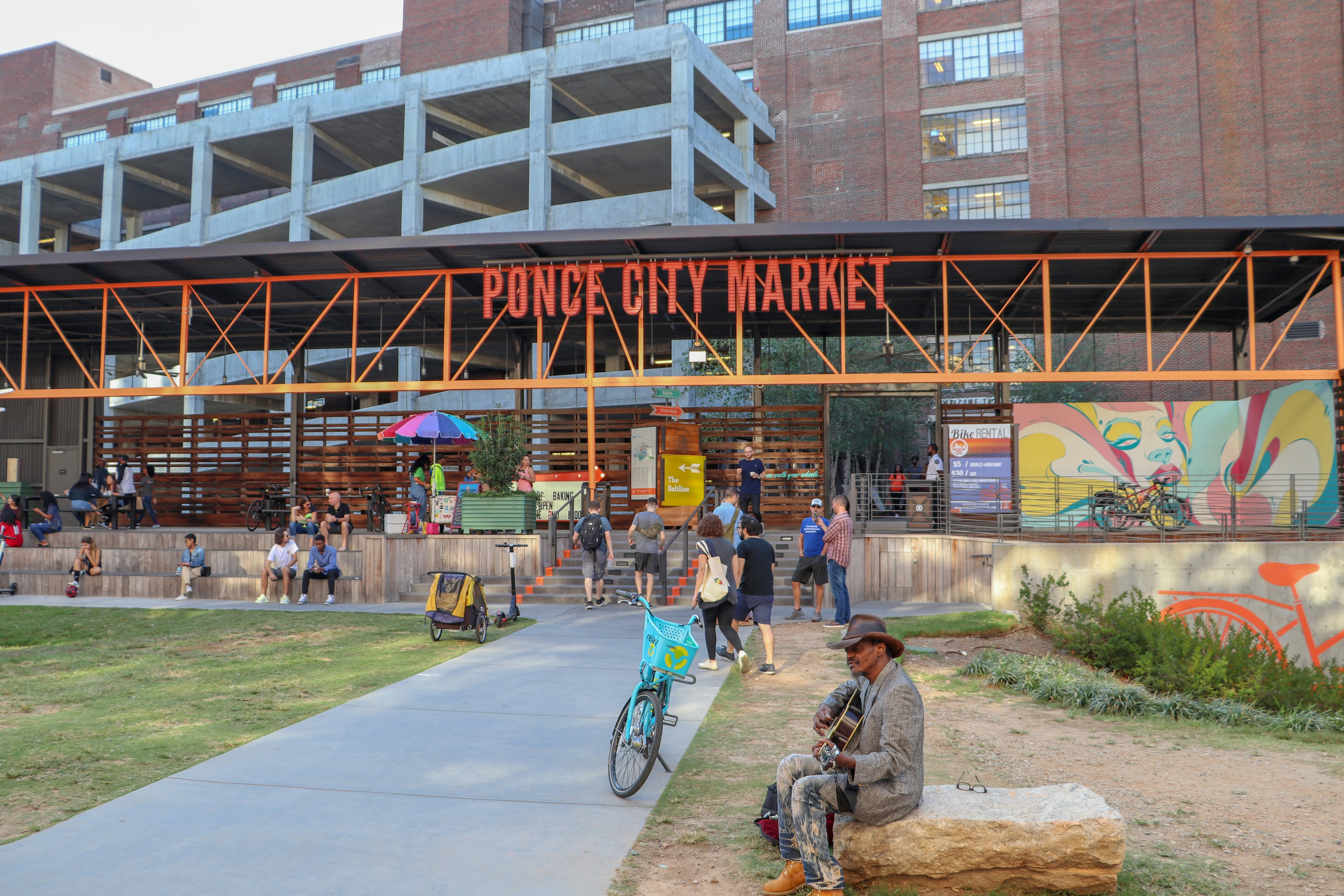 A musician with a guitar sits in a grassy area outside a public city market.