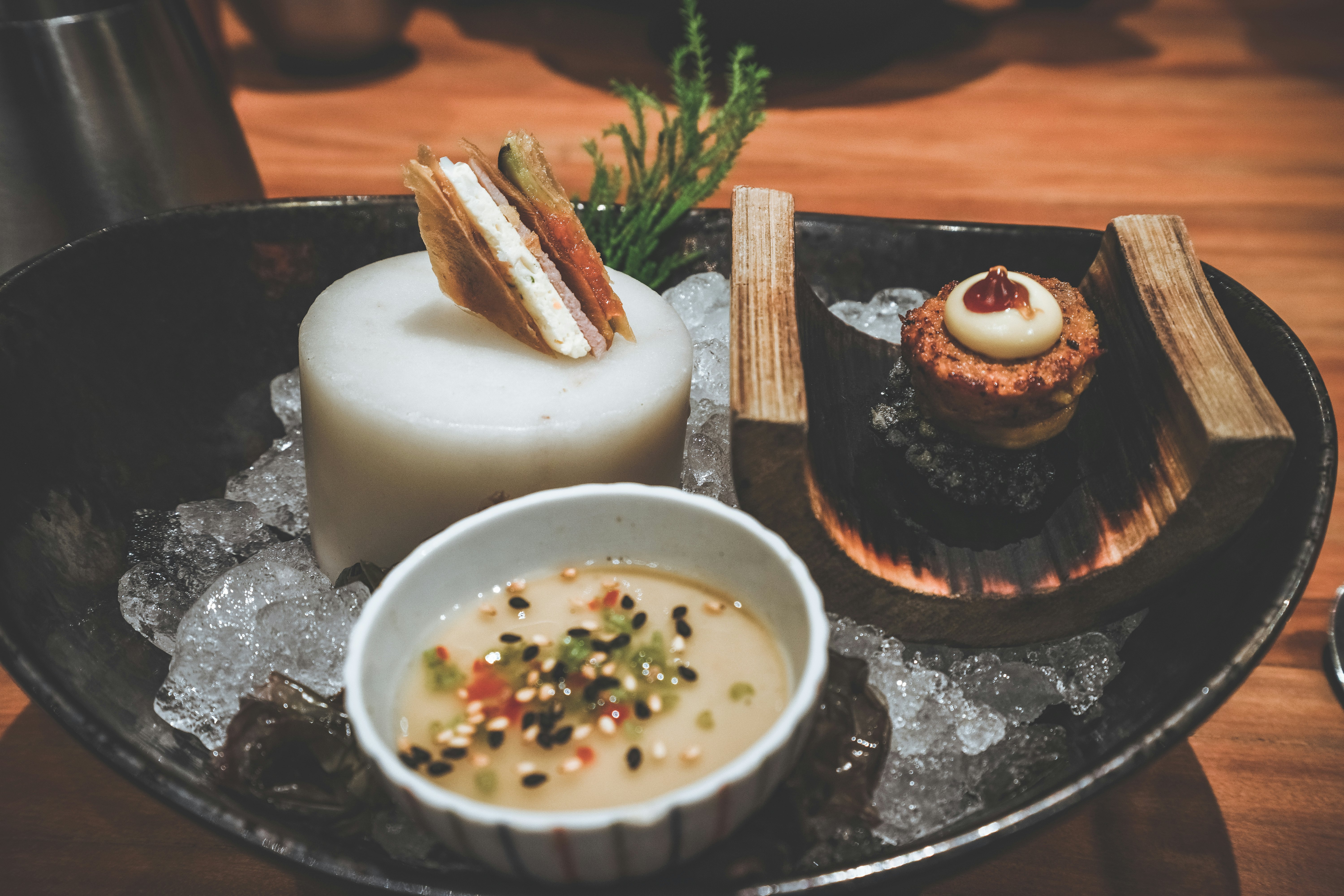 Three small plates of innovative food tucked among ice on a larger, black plate placed on a wooden table.