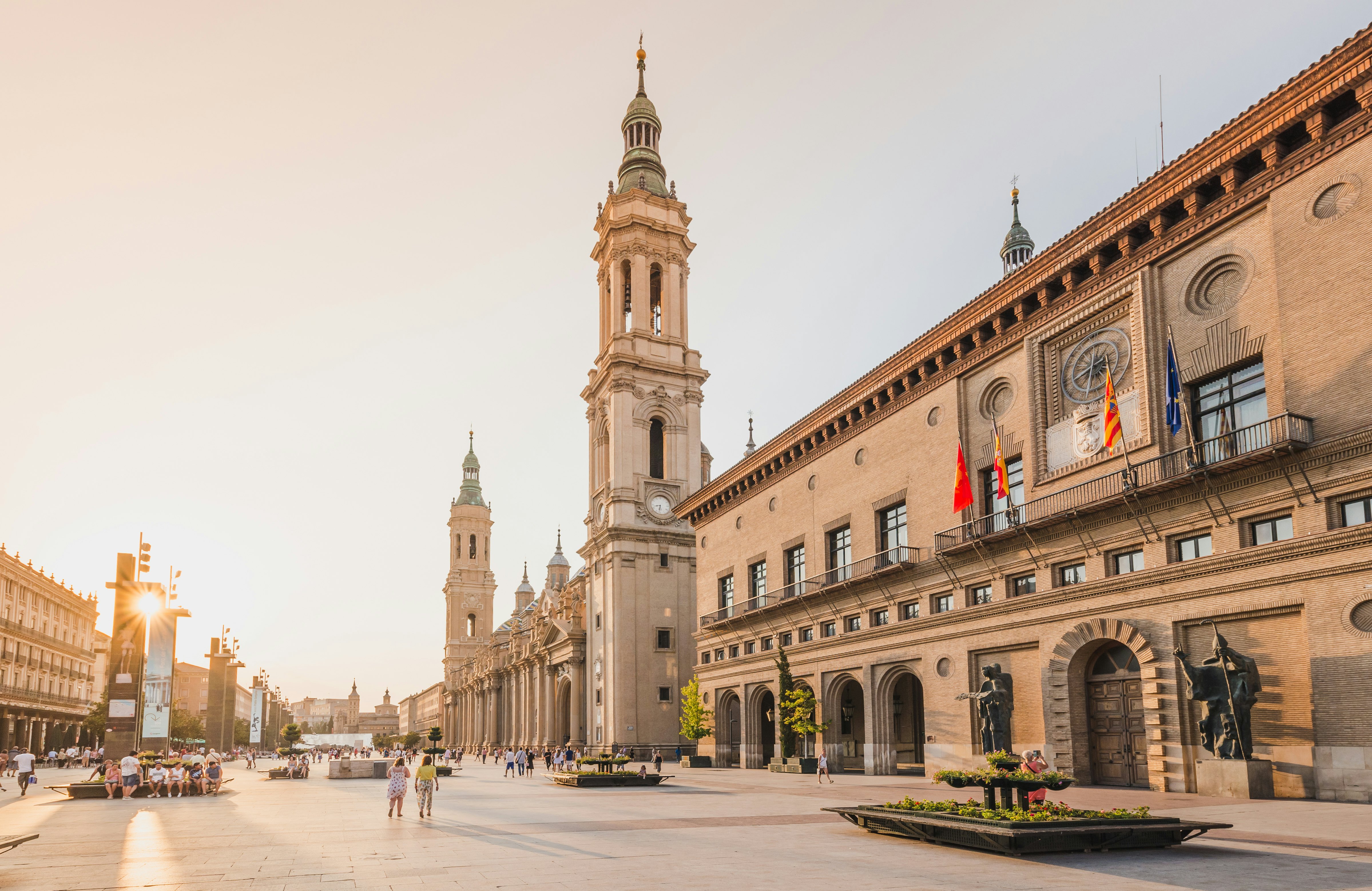 People wander thorugh a city square at sunset, with large administrative buildings to one side with two tall bell towers.