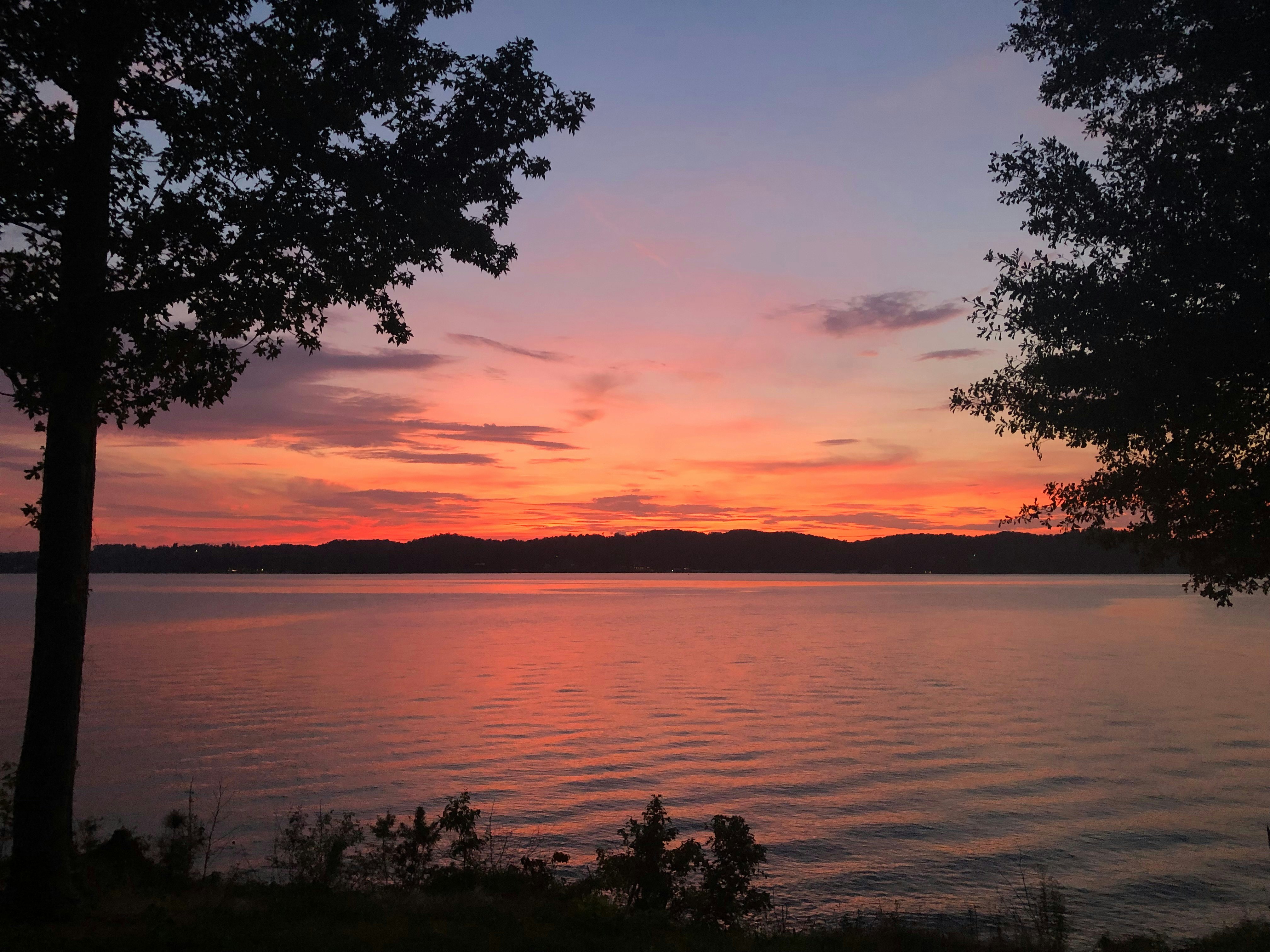 An orangey-pink sunset over a lake with hills silhouetted in the distance and trees in the foreground on either side.