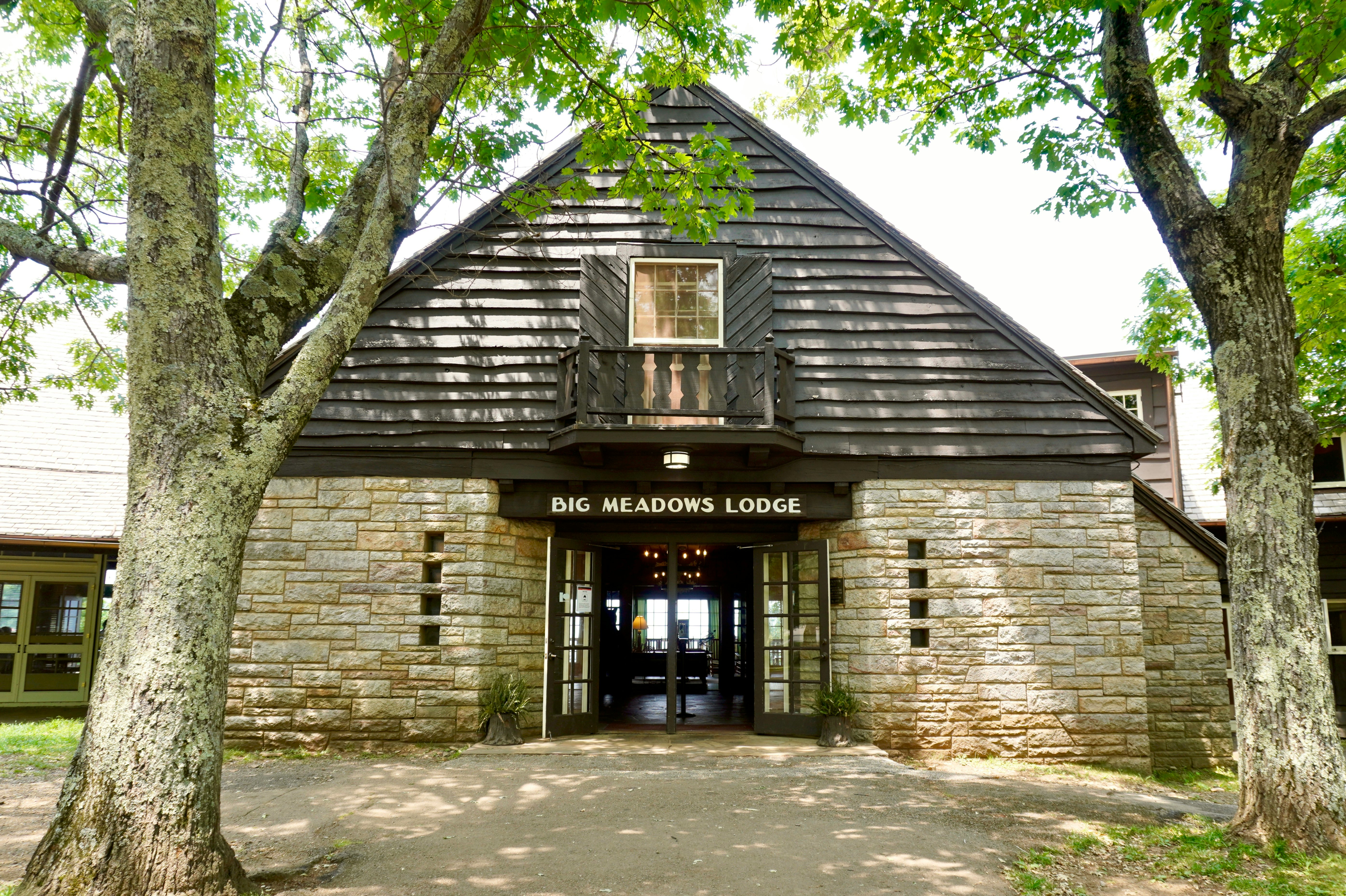 Photo of stone lodge with clapboard siding with a brown side above open doors reading "Big Meadows Lodge" in white capital letter. Sunlight casts shadows through deciduous trees onto a dirt walkway in the foreground.