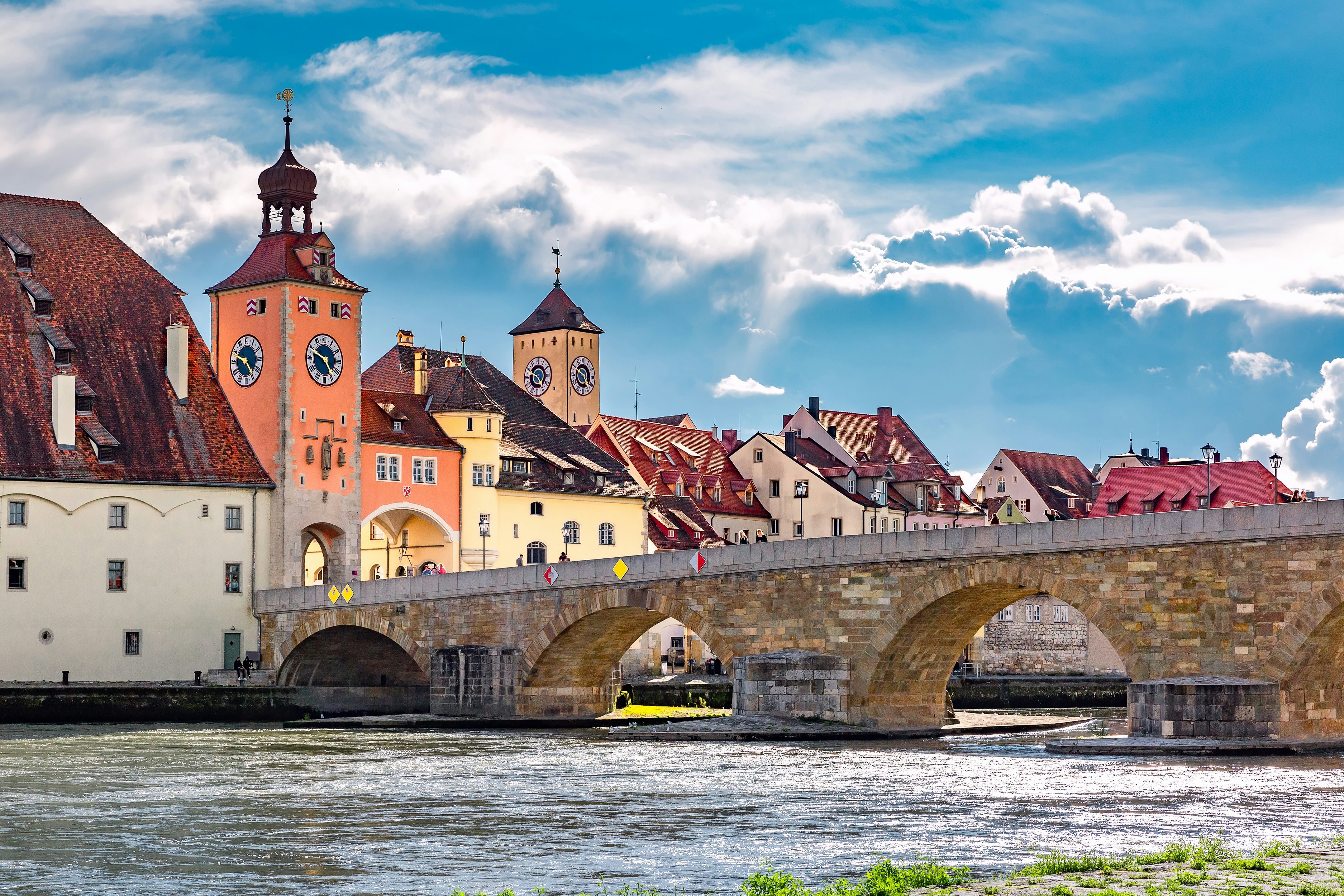 A stone bridge with several piers spans a river. Historic buildings are seen along the riverfront.