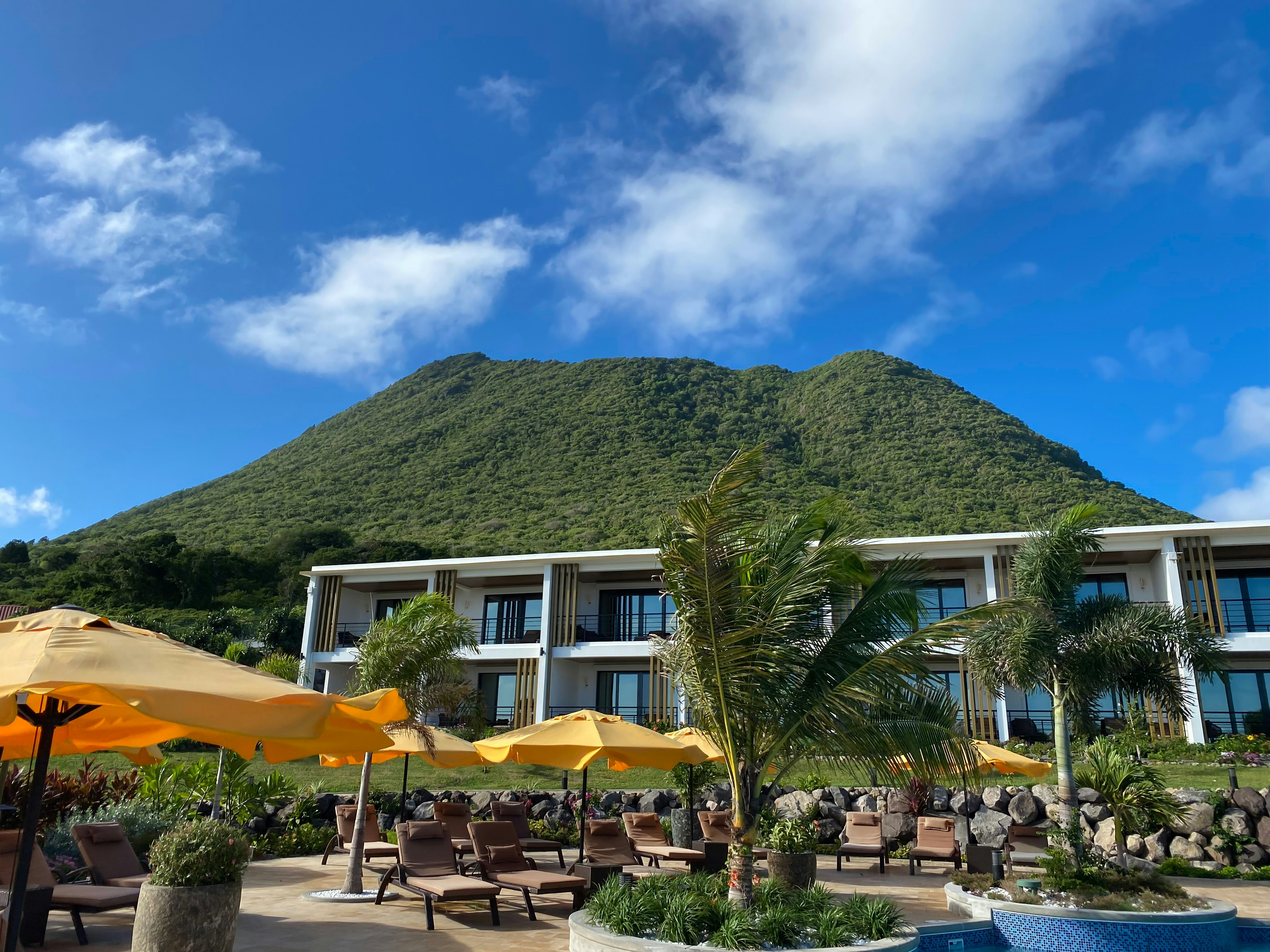 A hotel terrace, in the shadow of a large volcano, with sunloungers under yellow umbrellas.