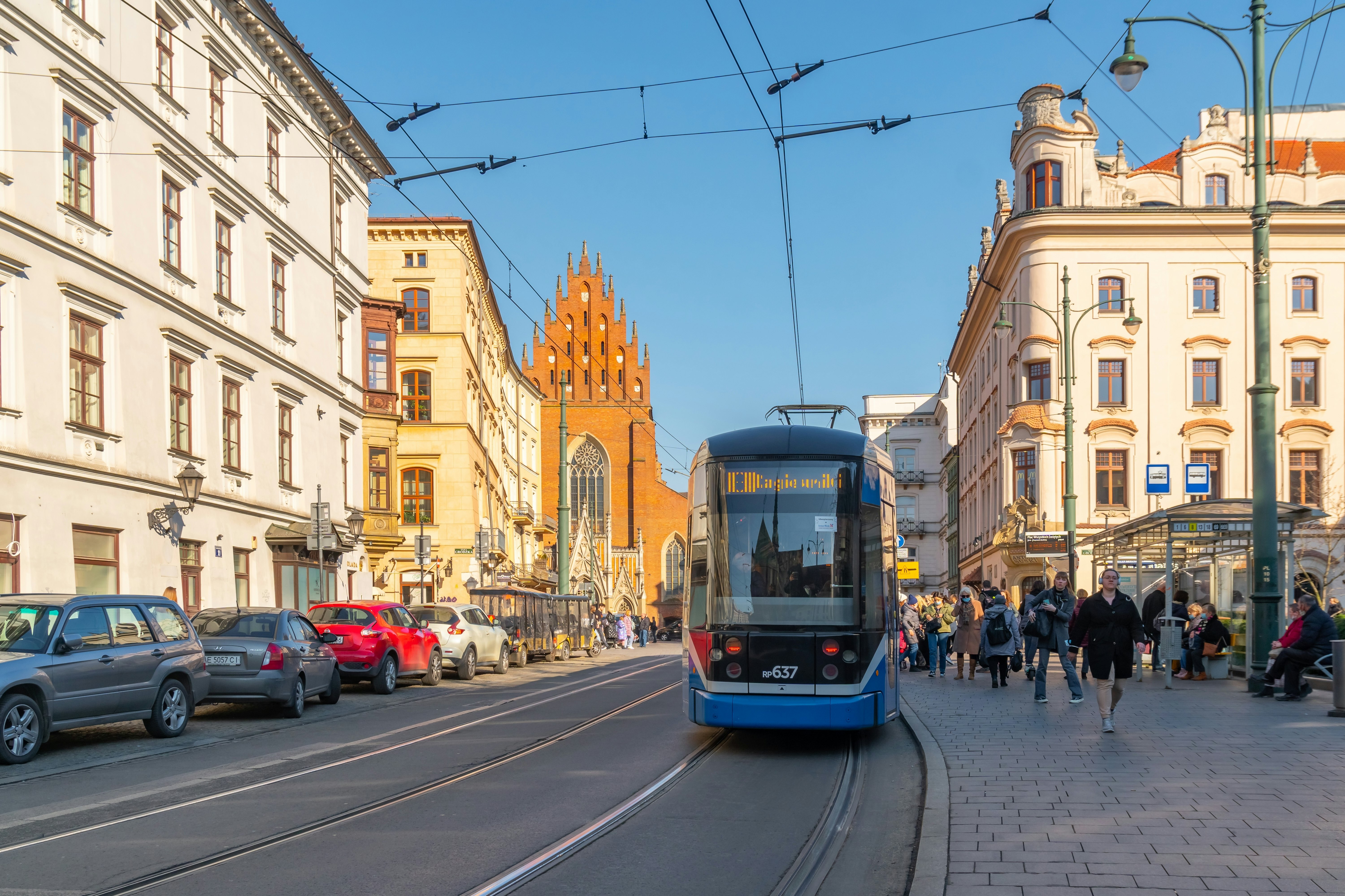 A blue tram passing a stop for passengers in °°ù²¹°ìó·É; a church is visible at the end of the roadway.