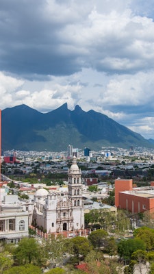 A view of a Spanish Colonial church surrounded by green trees a large city in the background with a mountain in the distance