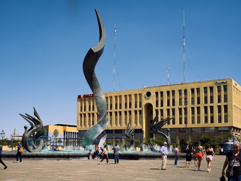 A sculptural fountain sits in a big square in front of a rectangle yellow building