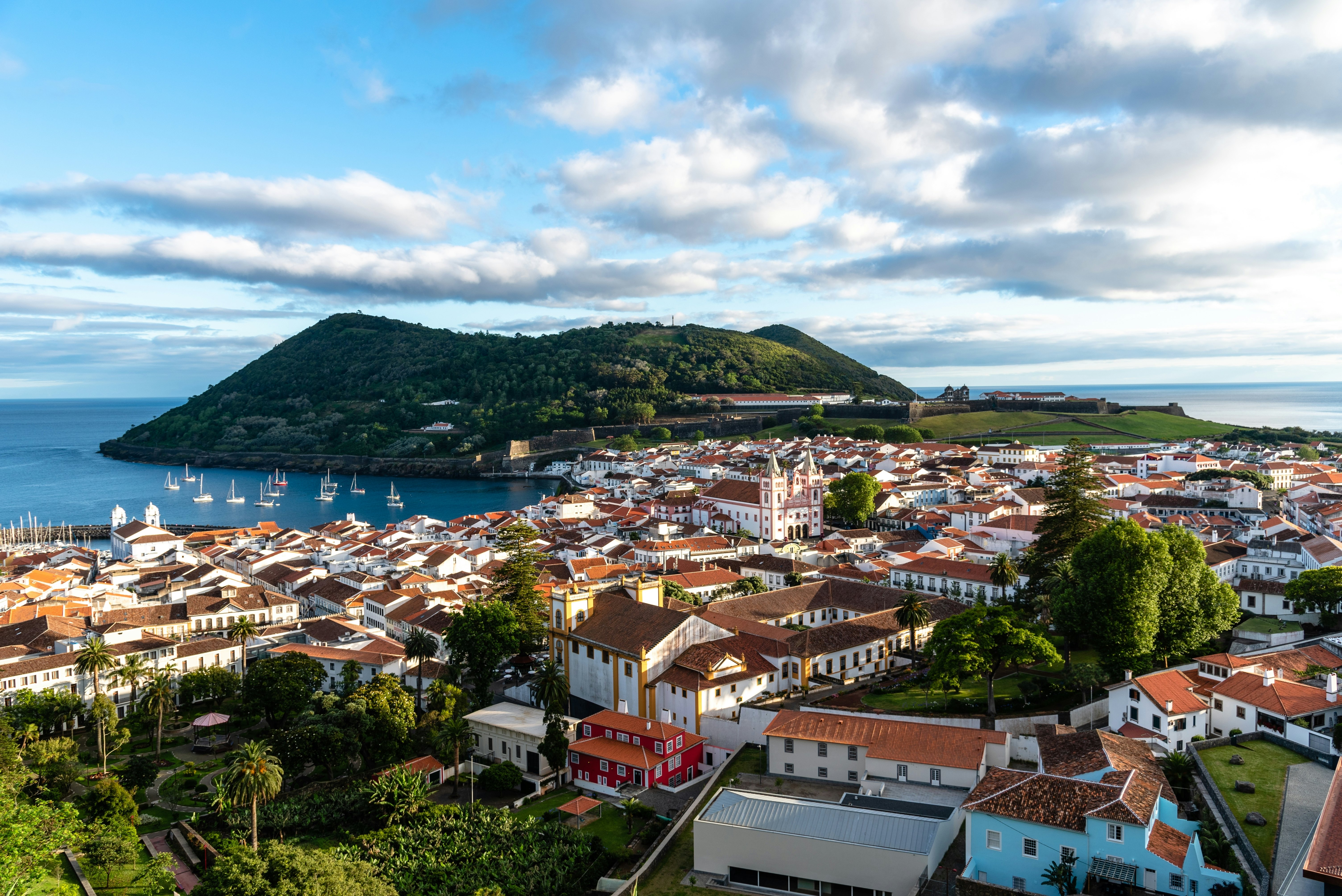 Aerial view of town of mostly white buildings with reddish roofs, with a bay and forested hillside in the distance on a sunny day.