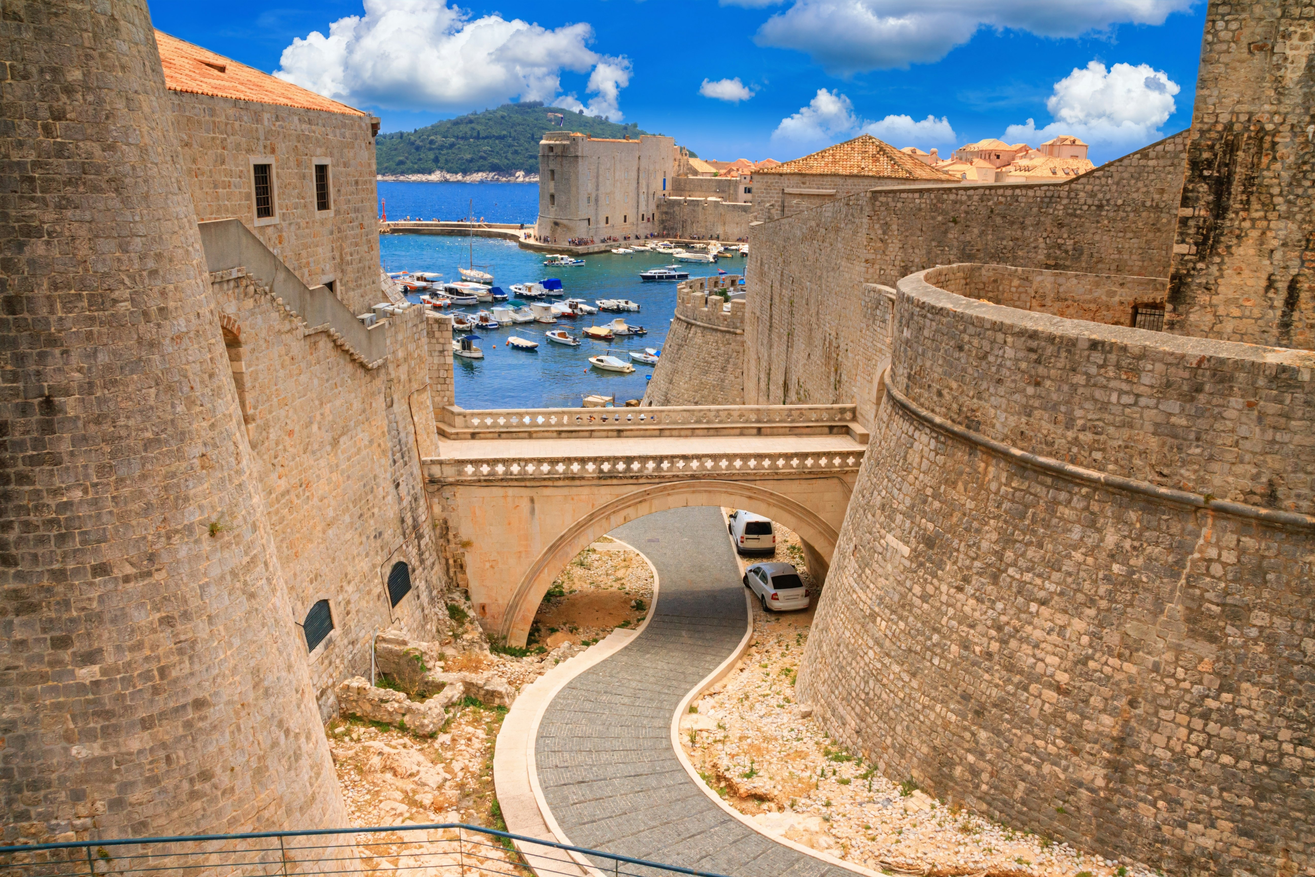A graded walkway runs down to blue water under a golden stone bridge connecting two parts of a golden brick fortress; two cars are parked under the bridge, and there are boats in the water.