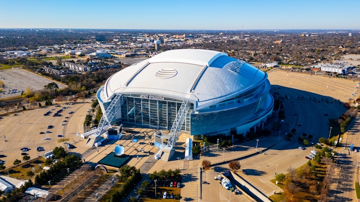 Round stadium amidst a large parking lot with a city stretching all around it