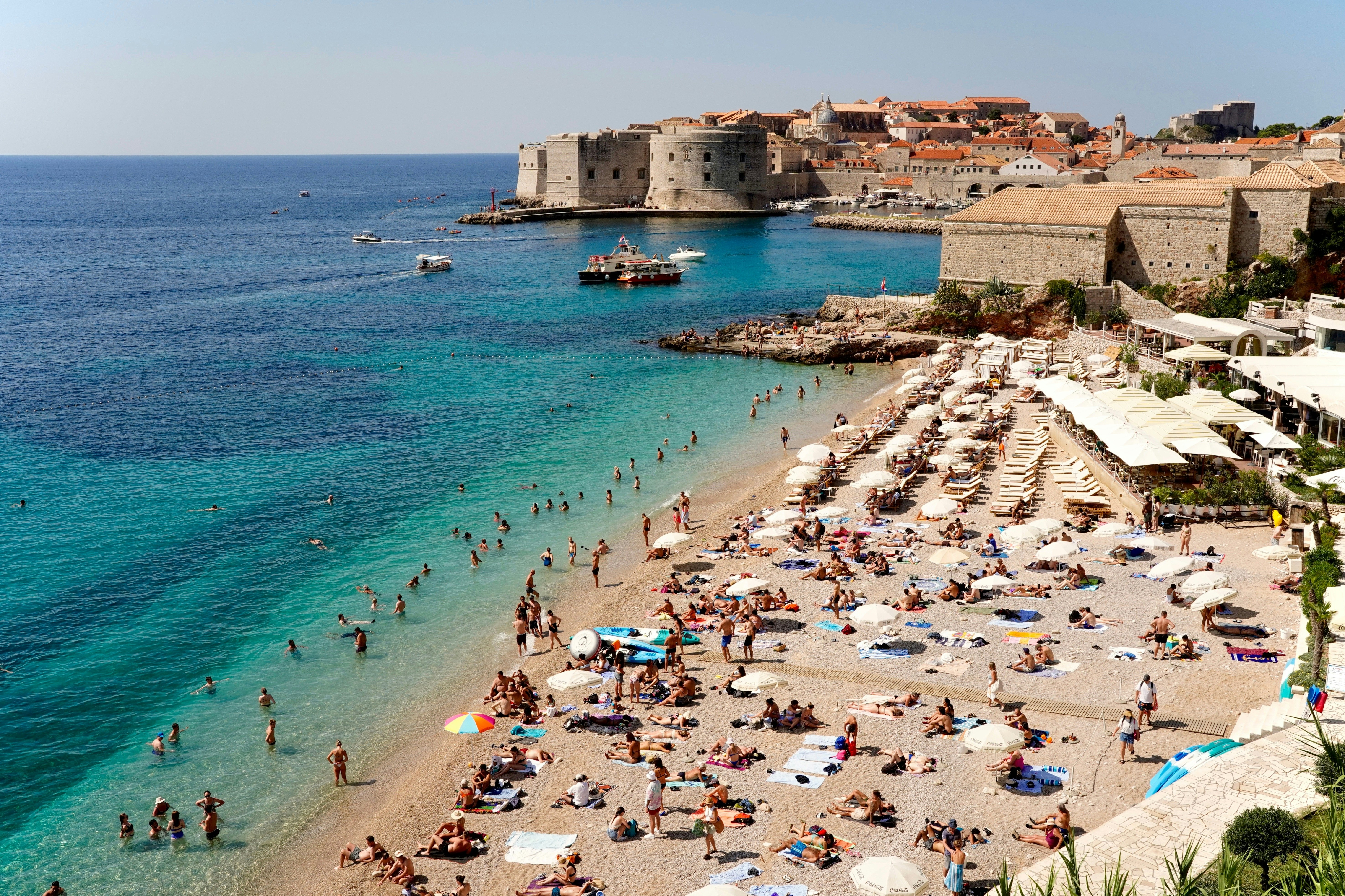 A crowded sandy beach by turquoise water; a fortress at the edge of Dubrovnik is in the background.