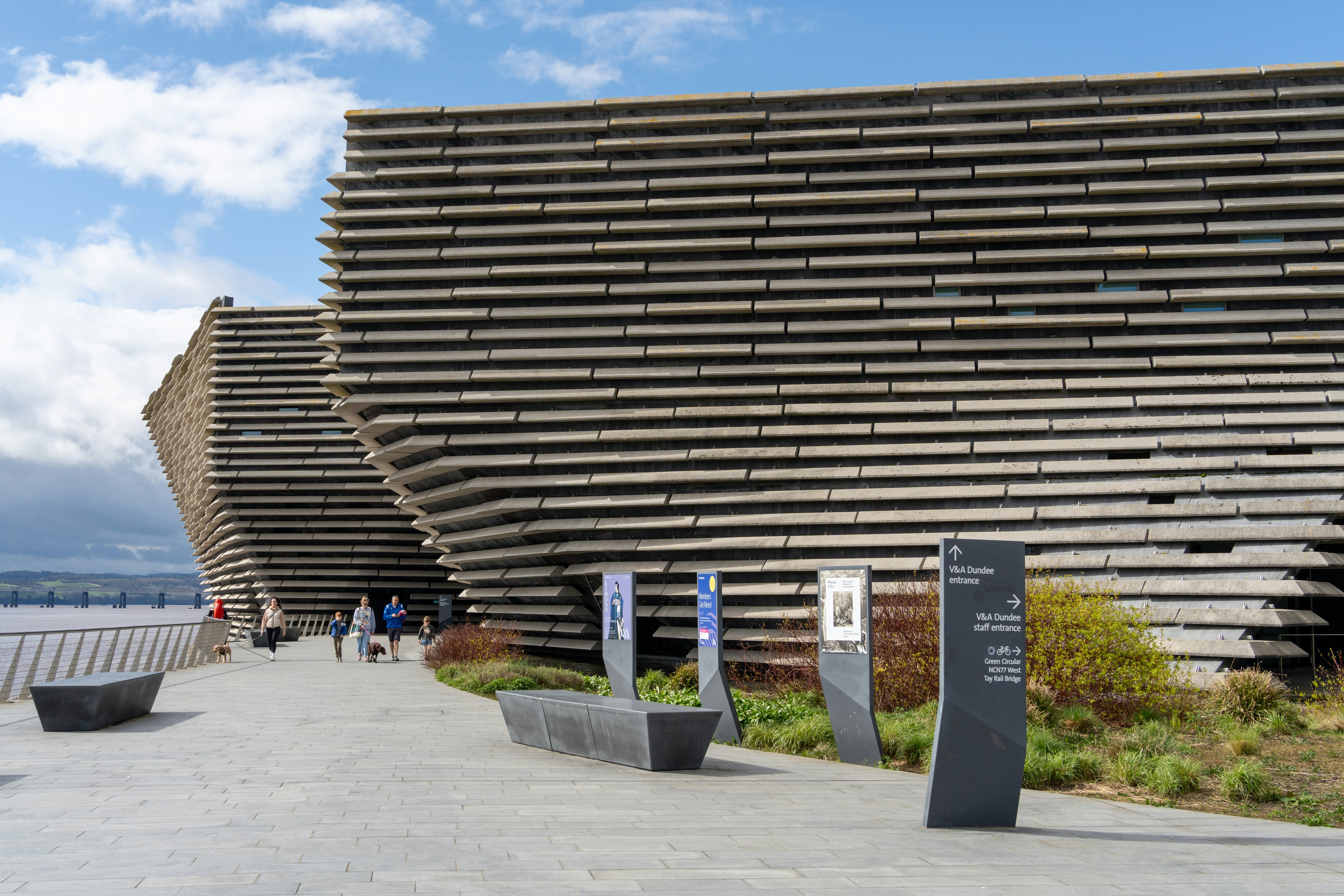 Modern buildings on an esplanade with concrete walkway, directional signs and pedestrians in the distance on a partly sunny day.