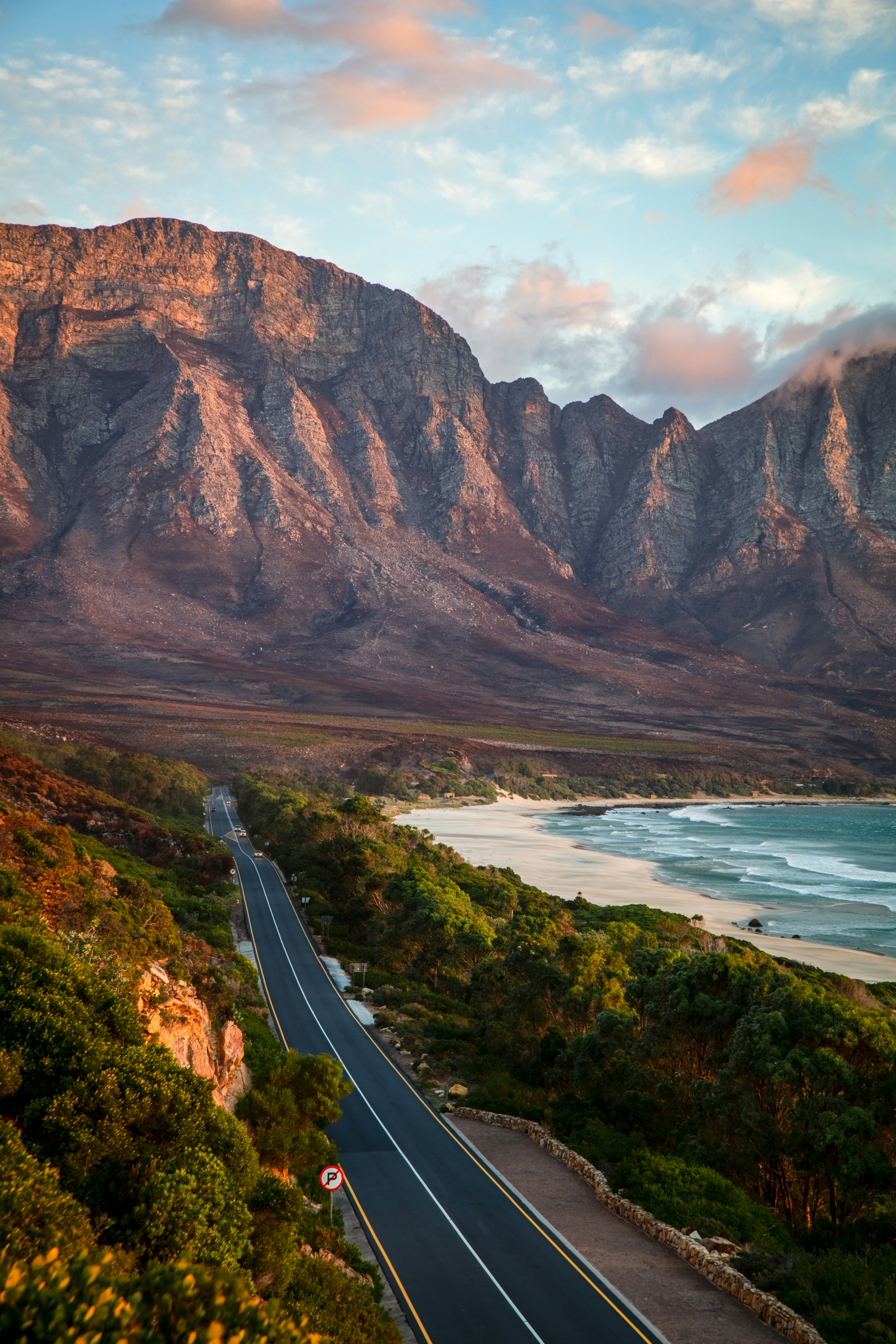 Two-lane road surrounded by green trees with ocean to the right and reddish rocky mountains in the distance at sunset.