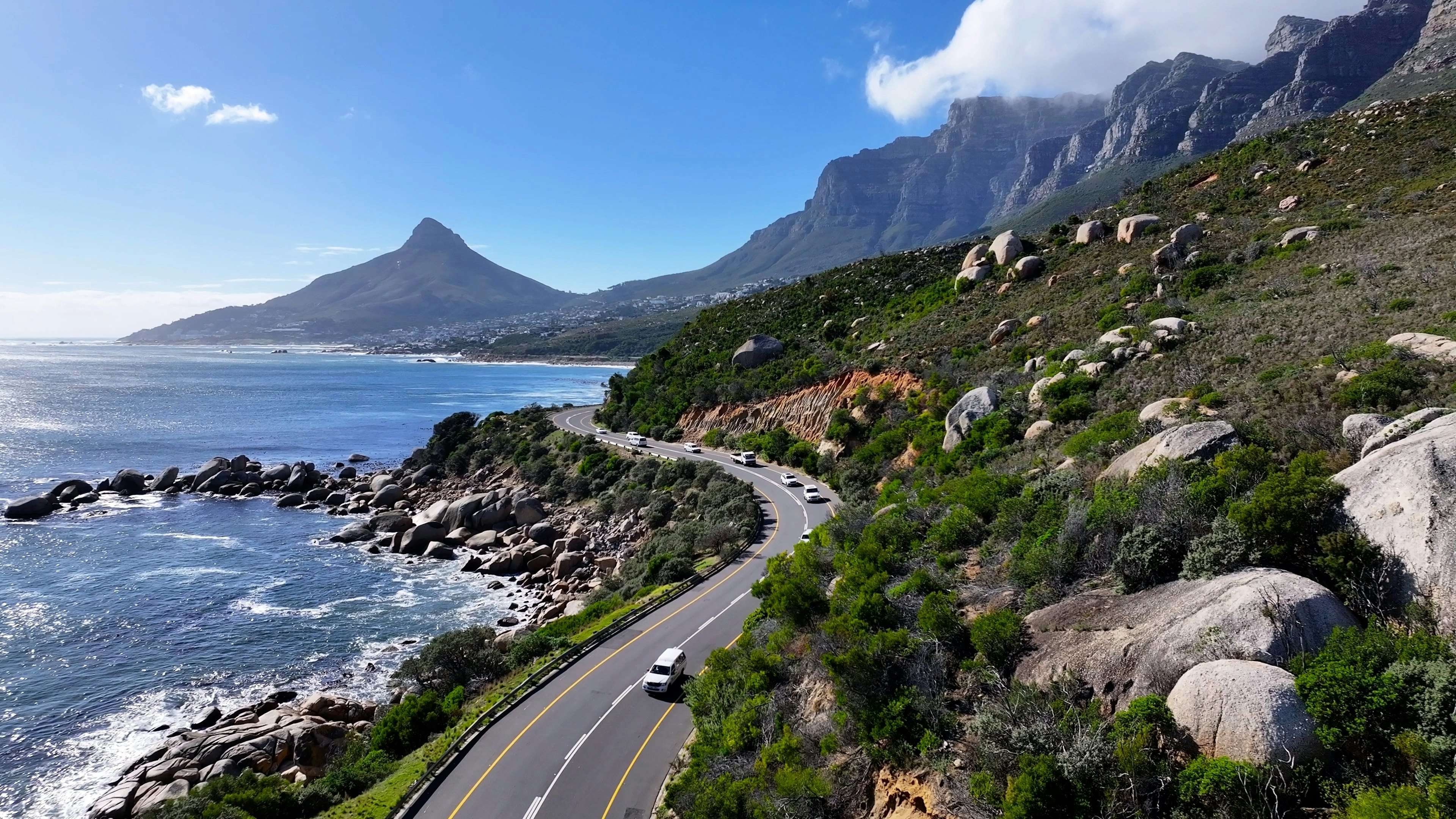 Cars, taxis and minibuses drive on a coastal road between ocean and a mountain.