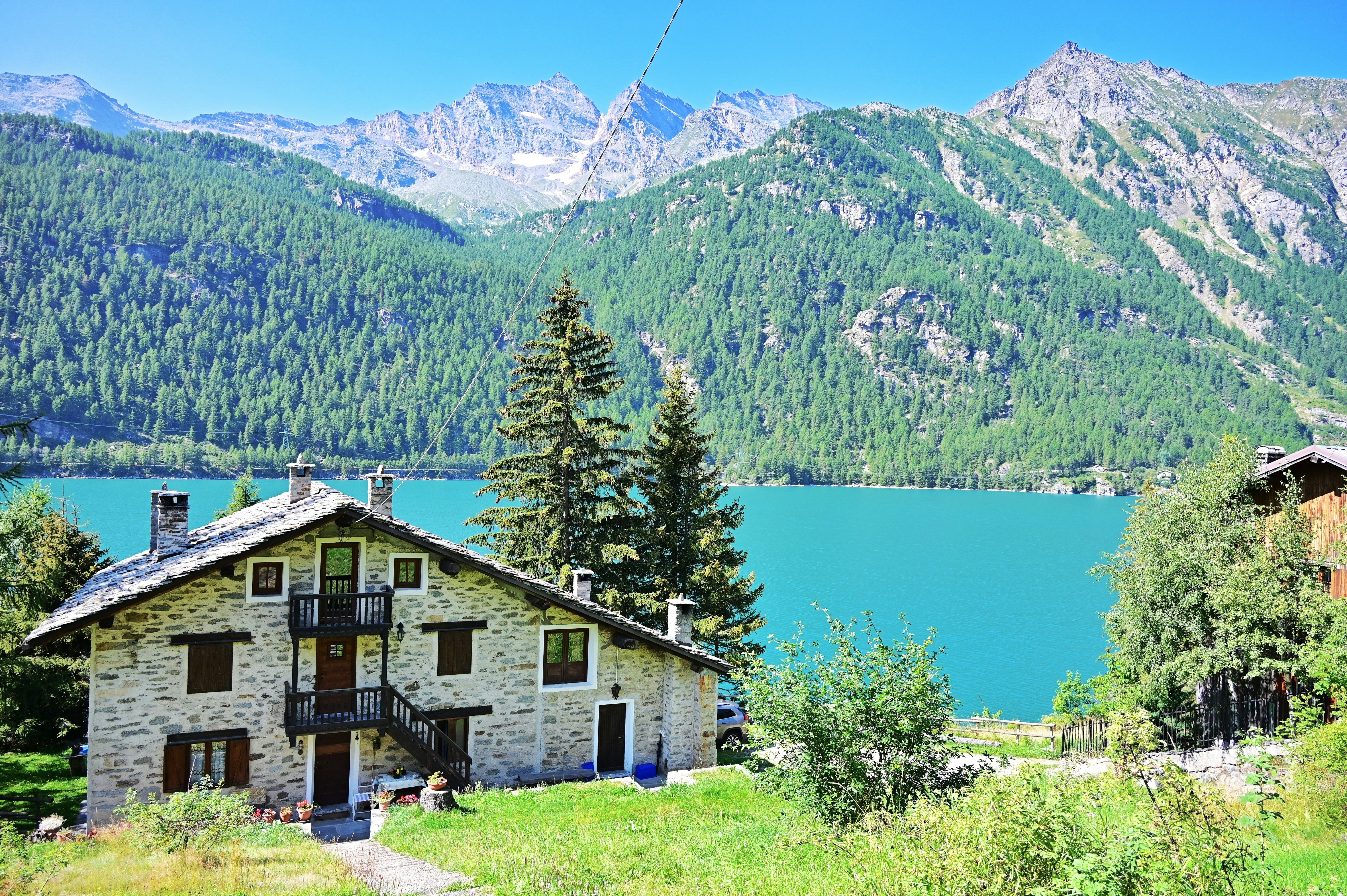 A chalet-style house is pictured on the edge of a lake, with mountains rising on the other shore.