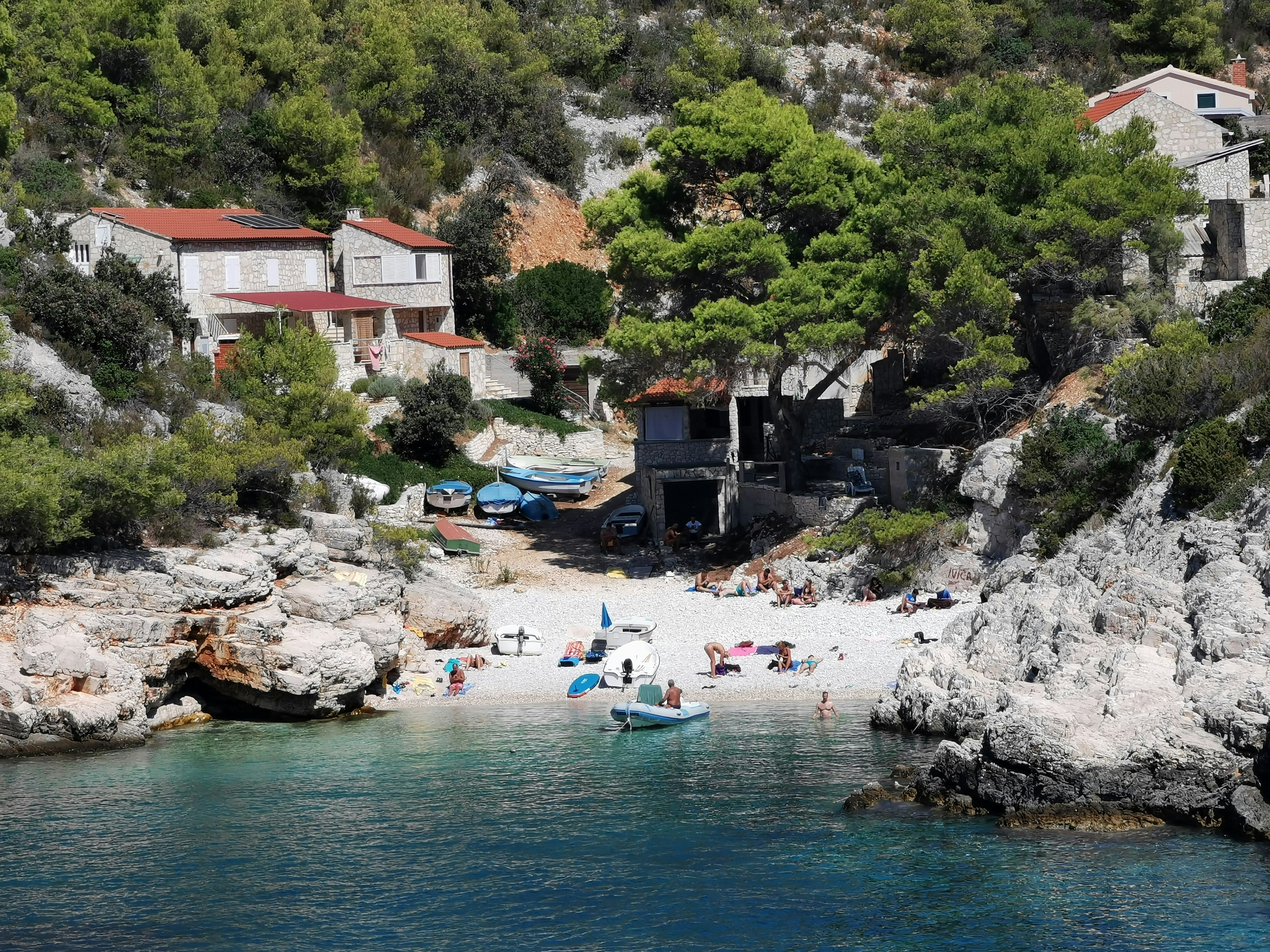 Stone buildings with red roofs surround a small inlet where boats are pulled up on shore and people sunbathe and swim in the turquoise sea.