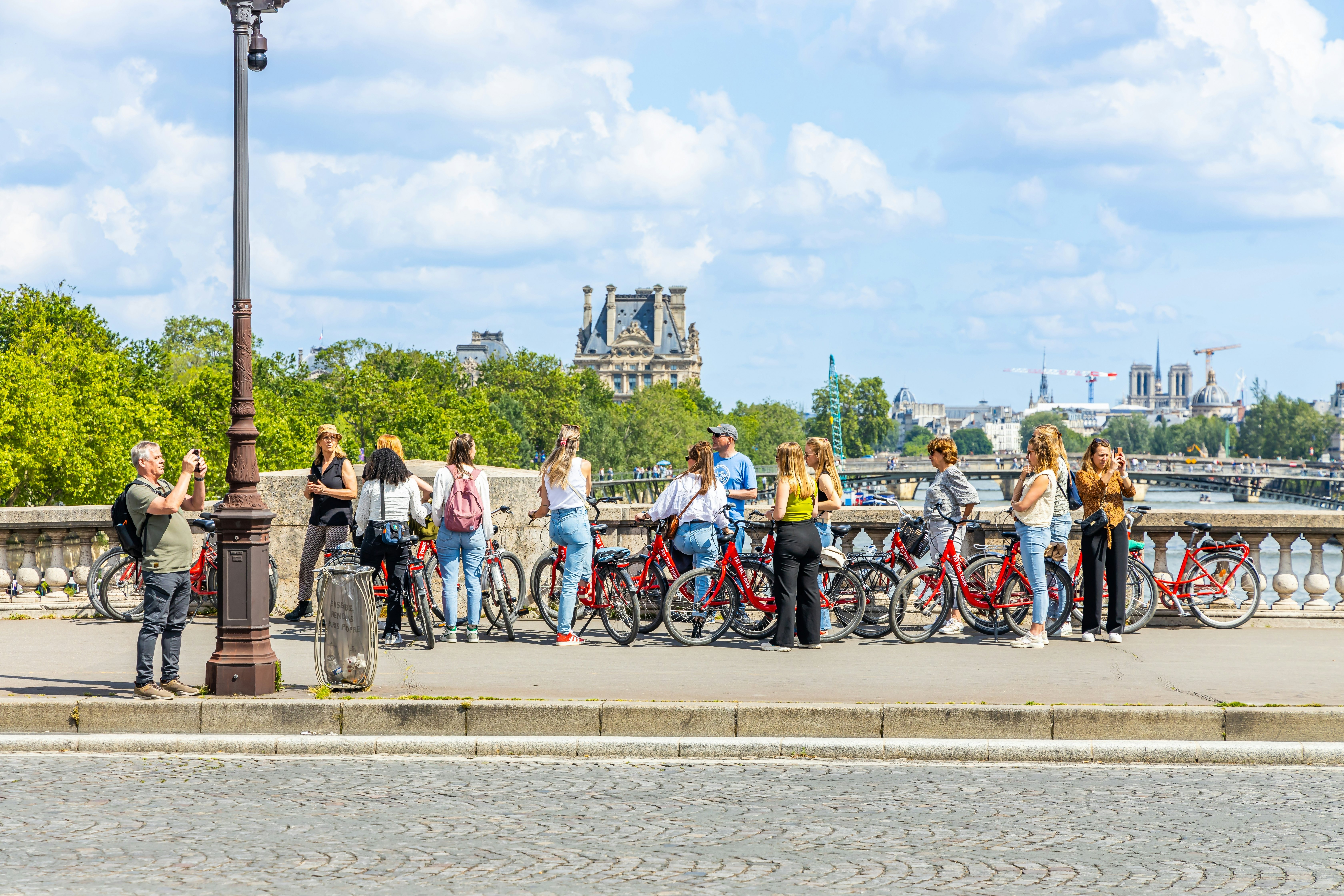 A group of people, each with a red bike, listen to a tour guide while pausing on a bridge over a river in a city.