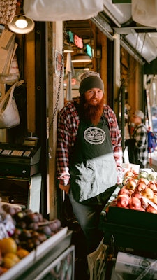 A vendor with a long red beard stands behind a fresh fruit stand