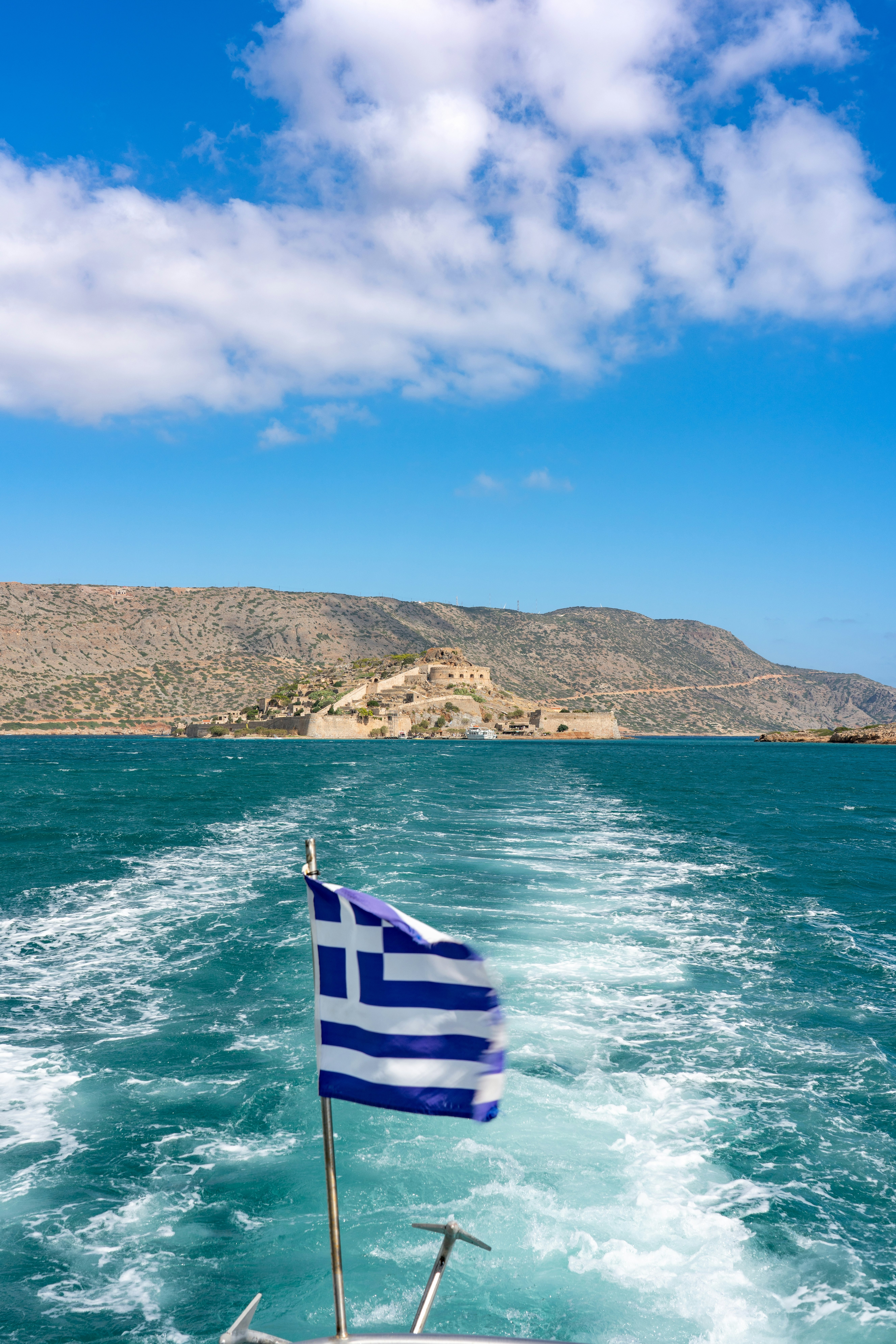 A vibrant Greek flag flutters on a boat approaching Spinalonga Island, Crete.