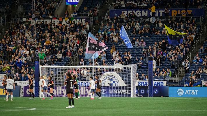 Fans fill the stands of a stadium while two teams of women play soccer in front of a goal