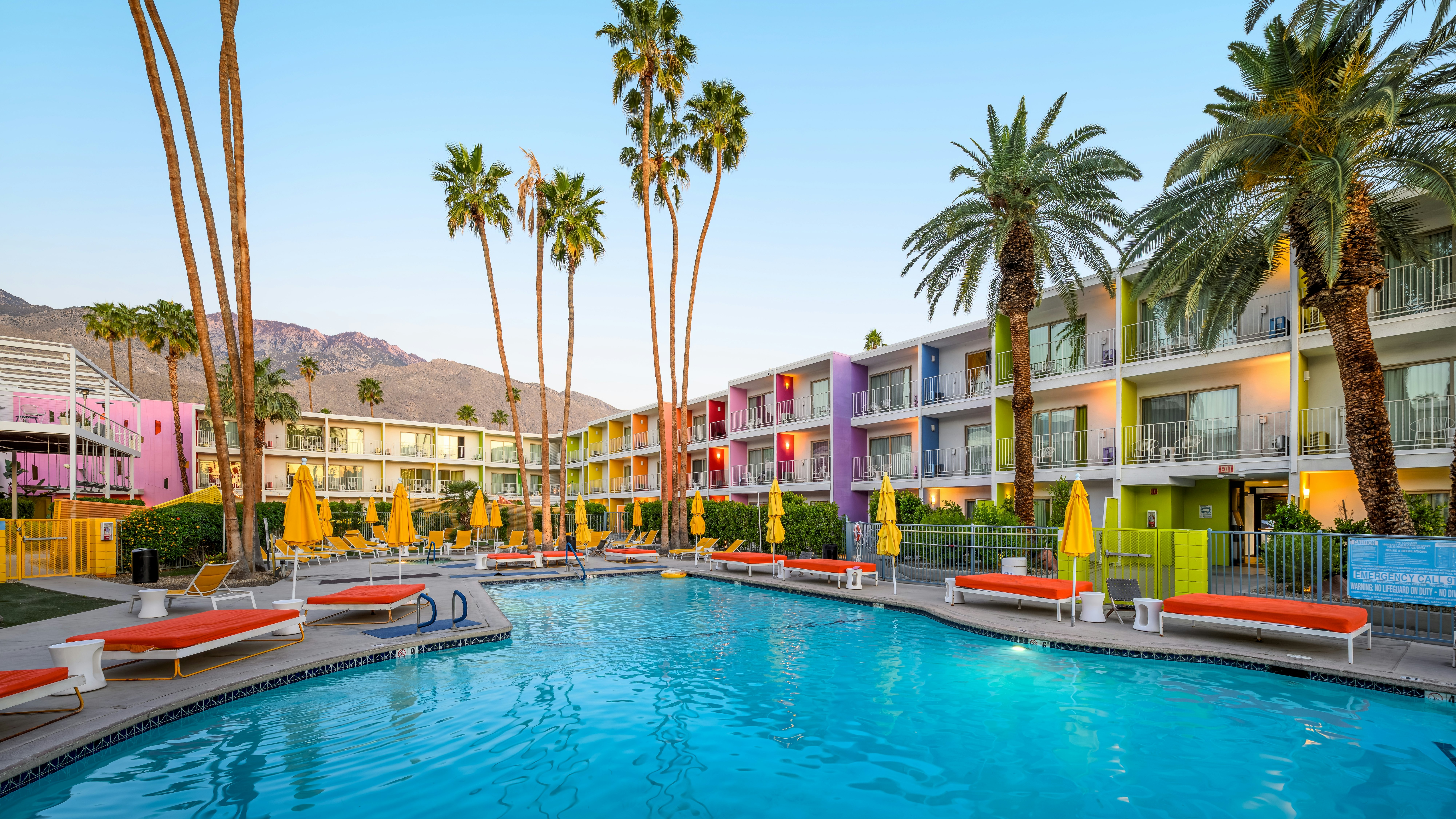 A pool surrounded by orange-cushioned benches, with closed yellow umbrellas, in the courtyard of a three-story hotel with colorful panels between the balconies.