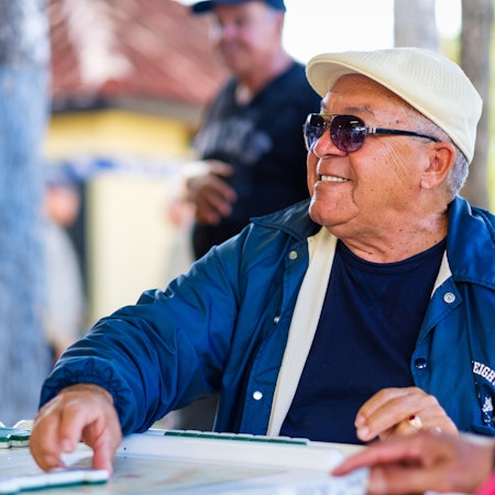 Man in a blue jacket and sunglasses plays dominos with a smile.