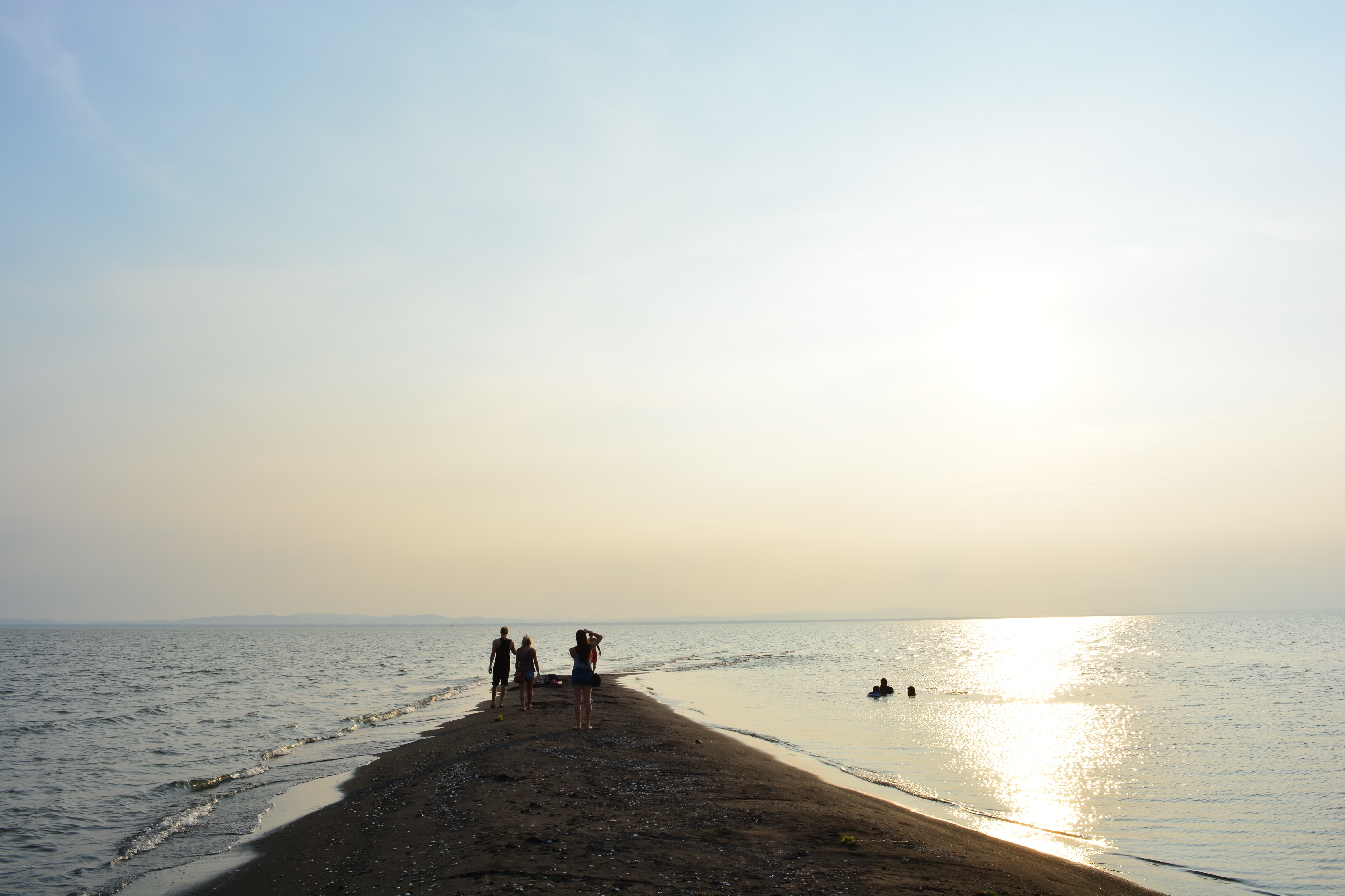 Five people on a narrow spit of sand surrounded by the ocean at sunset. Three people are swimming in the sea.