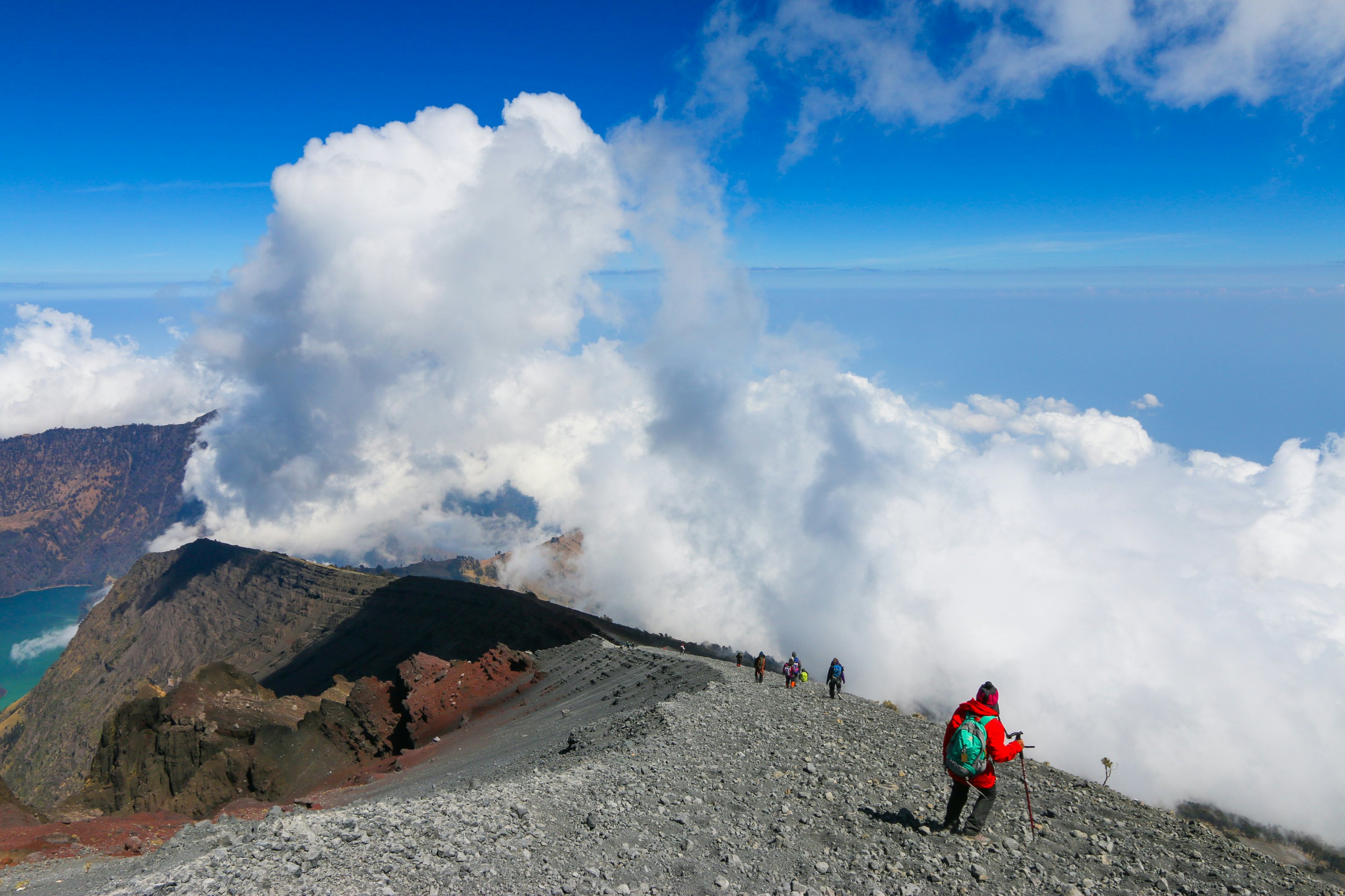 Hikers follow the gravelly ridge of a volcano summit. Clouds can be seen below the hikers, rising from the volcano crater.