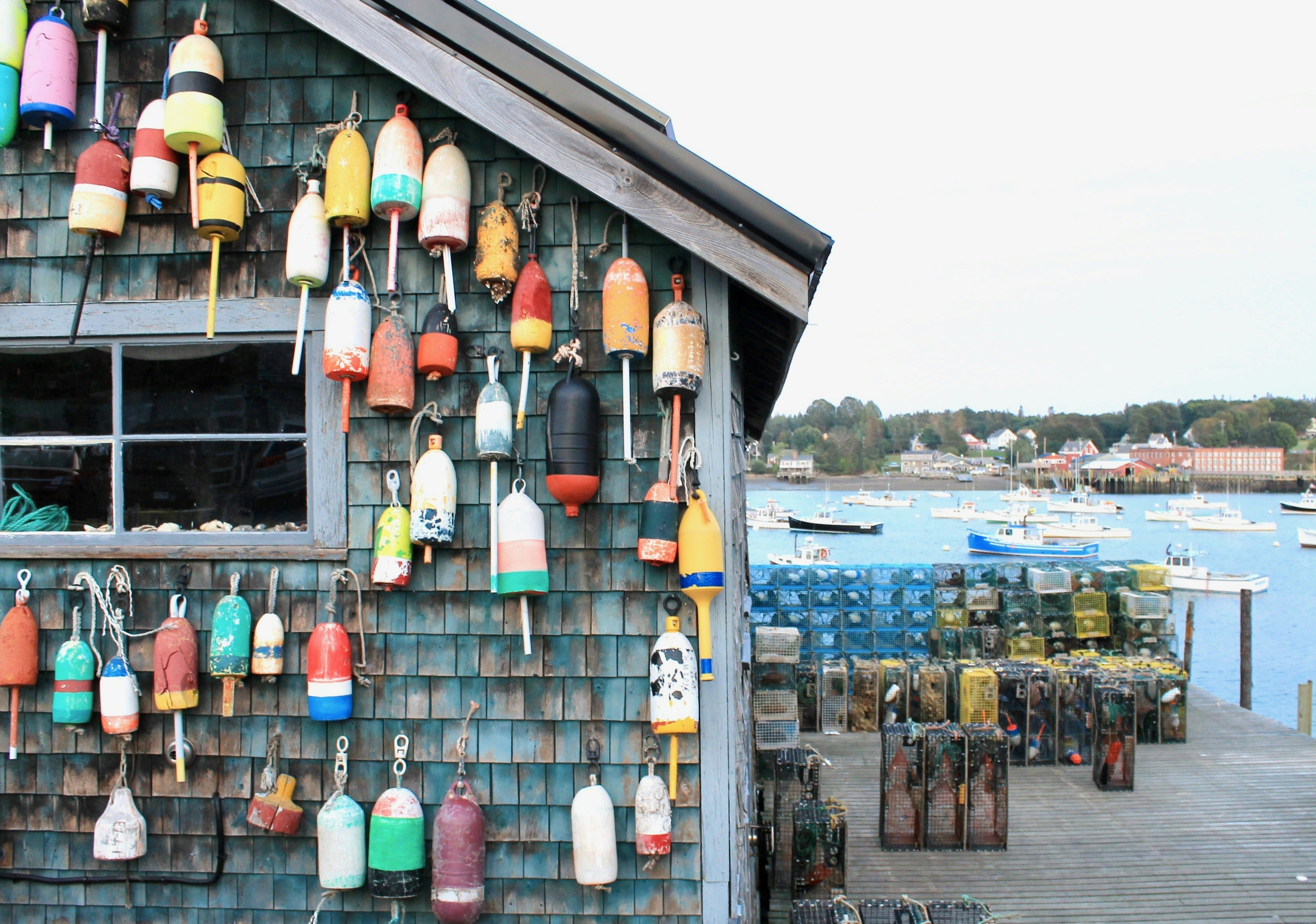 Shingled building covered in colorful buoys with stacks of lobster cages to the right on a dock, and boats on the water in the background