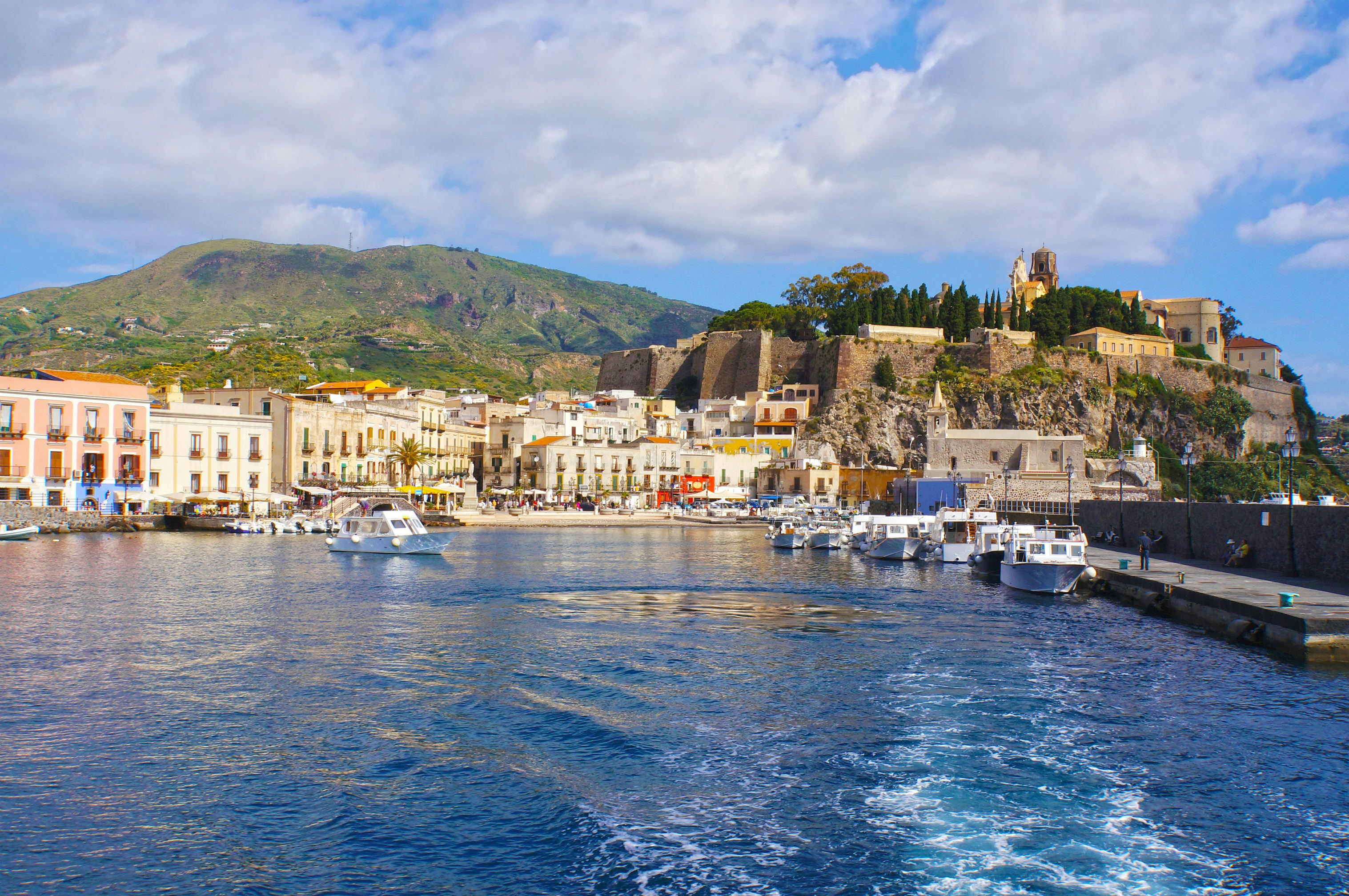 A small boat-filled marina with restaurants and shops lining the waterfront. Ancient city walls rise on a hillside.