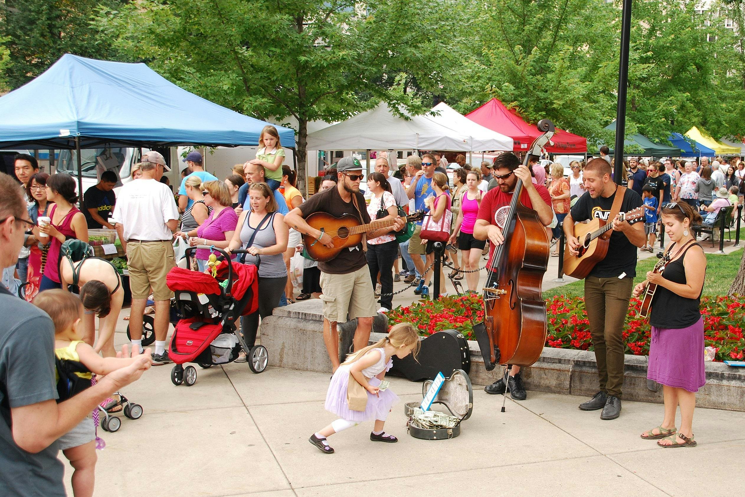 A crowded farmers market, with a four-person band playing as a child adds money to an open instrument case.